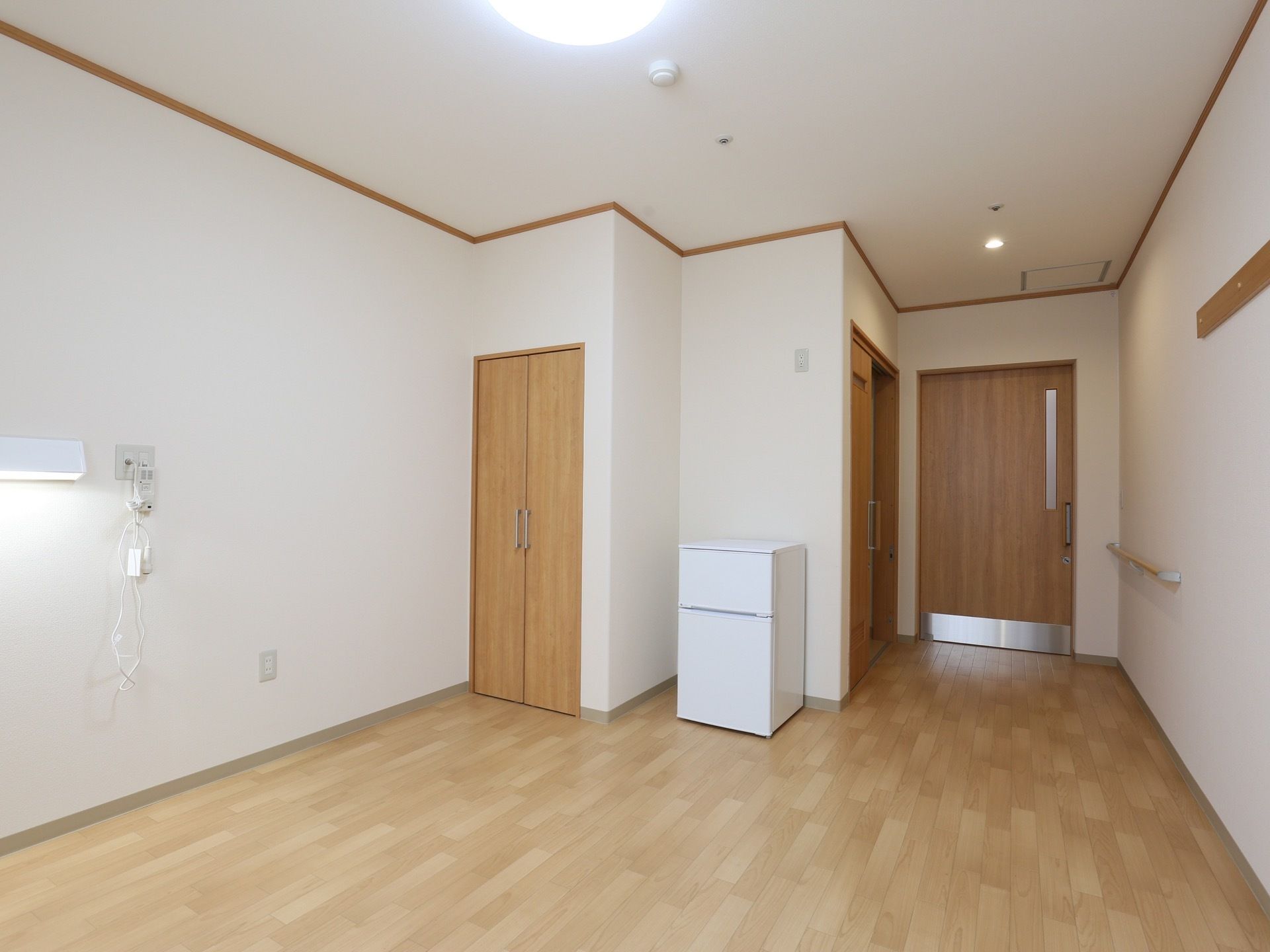 奥行きのあるフローリングの居室に2段扉の冷蔵庫が一台置かれている。収納が備え付けられていて、入り口近くの壁には手すりがある。