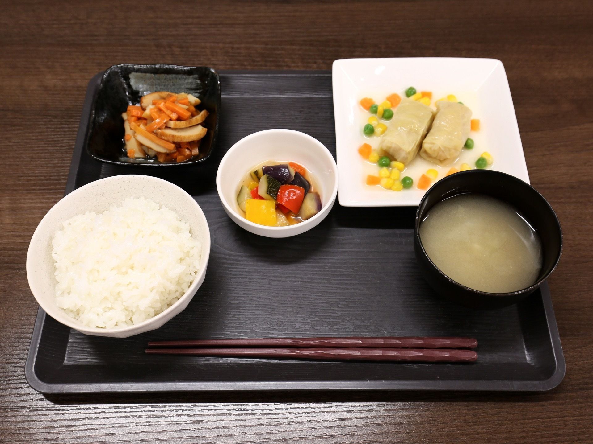 黒いお盆の上に、白米の茶碗、味噌汁、煮物の小鉢、ロールキャベツの白いお皿と炒め物の黒い小鉢が並べられている。