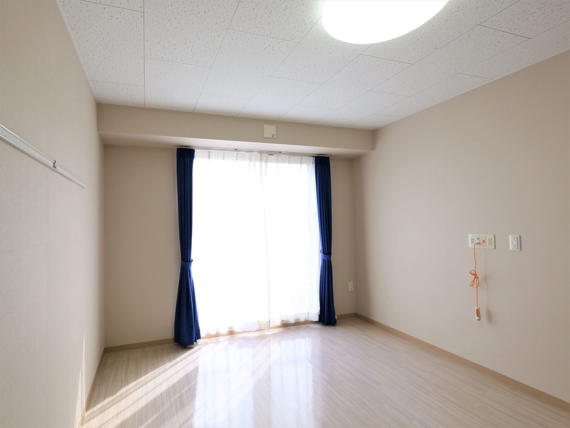 掃き出し窓があり、ブルーのカーテンが掛かっている。天井にはシーリングライトが設置され、壁にはナースコールが取り付けられている。
