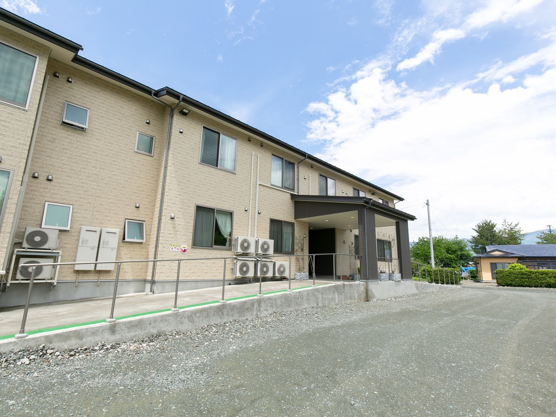 ベージュの外壁の2階建ての建物で玄関の上には大きなひさしが設けられている。外壁にエアコンの室外機が取りつけられている。
