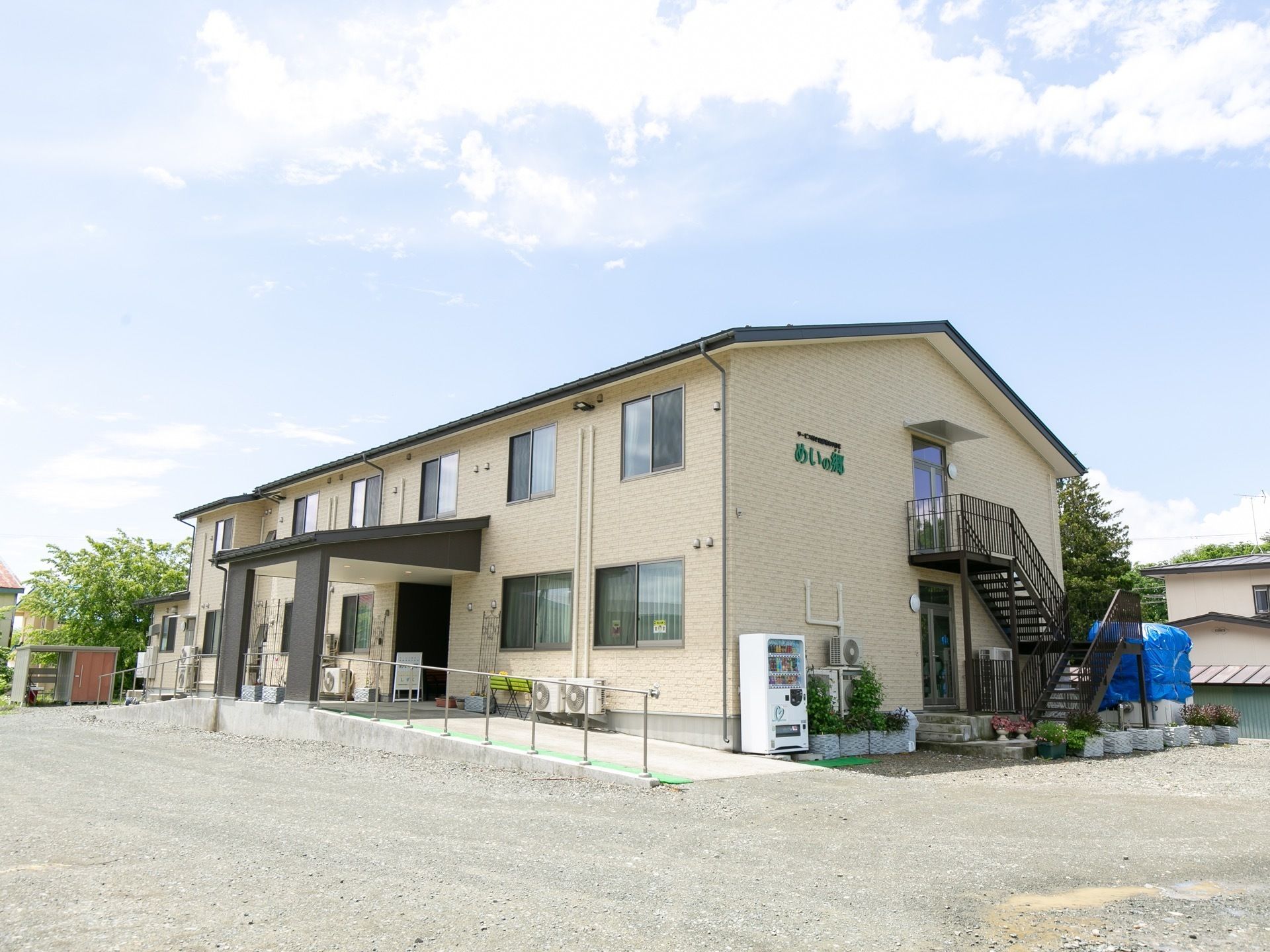建物の外壁に緑色の文字で施設名が書かれている。外階段が設置されており、その近くに自動販売機が置かれている。