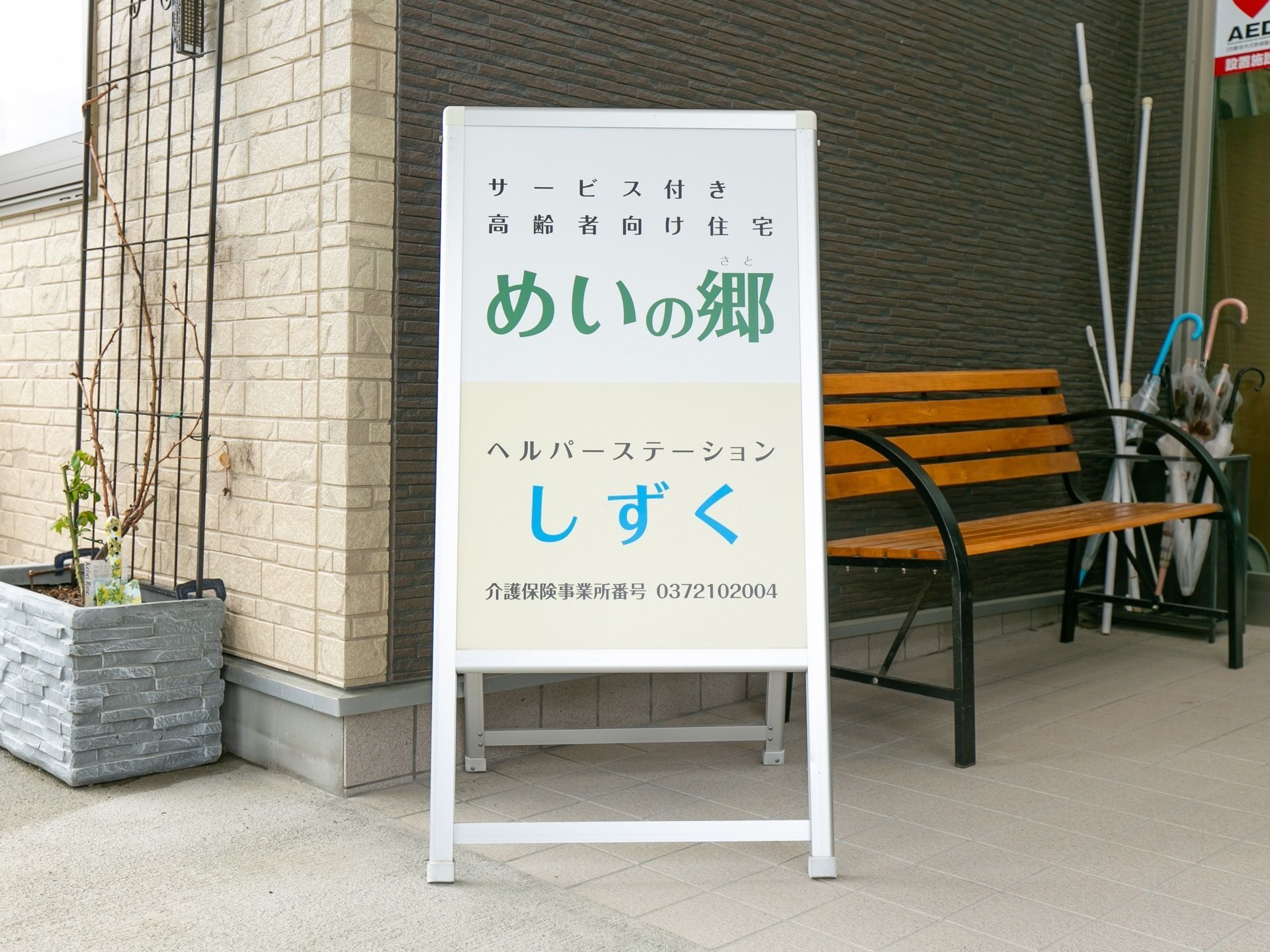 施設名が書かれた白い立看板があり、その右側に木製のベンチが置かれている。ベンチの奥には傘立てがある。