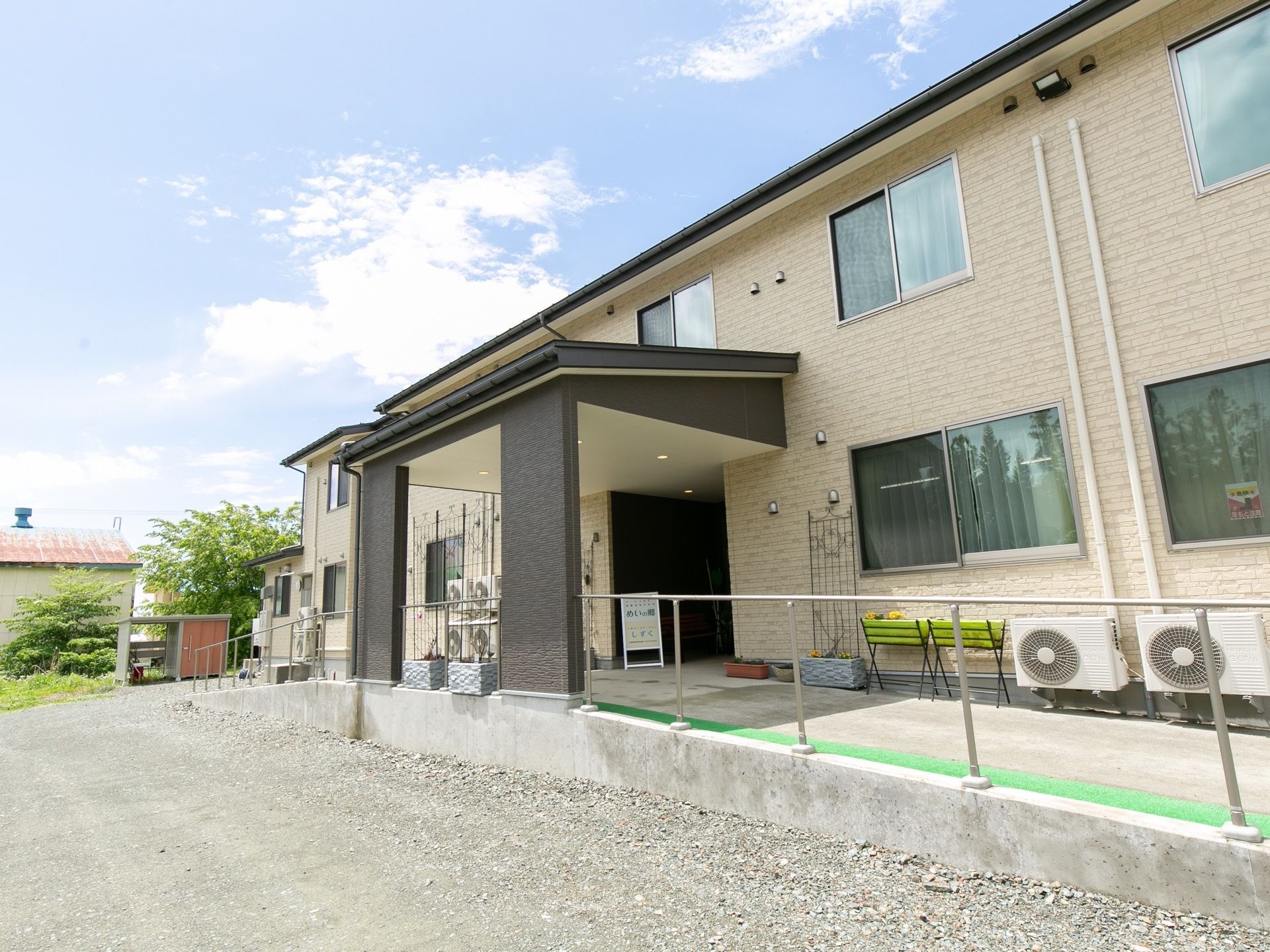 ベージュ色の外壁で落ち着いた雰囲気の建物である。玄関までの通路には手すり付きのスロープが設置されている。