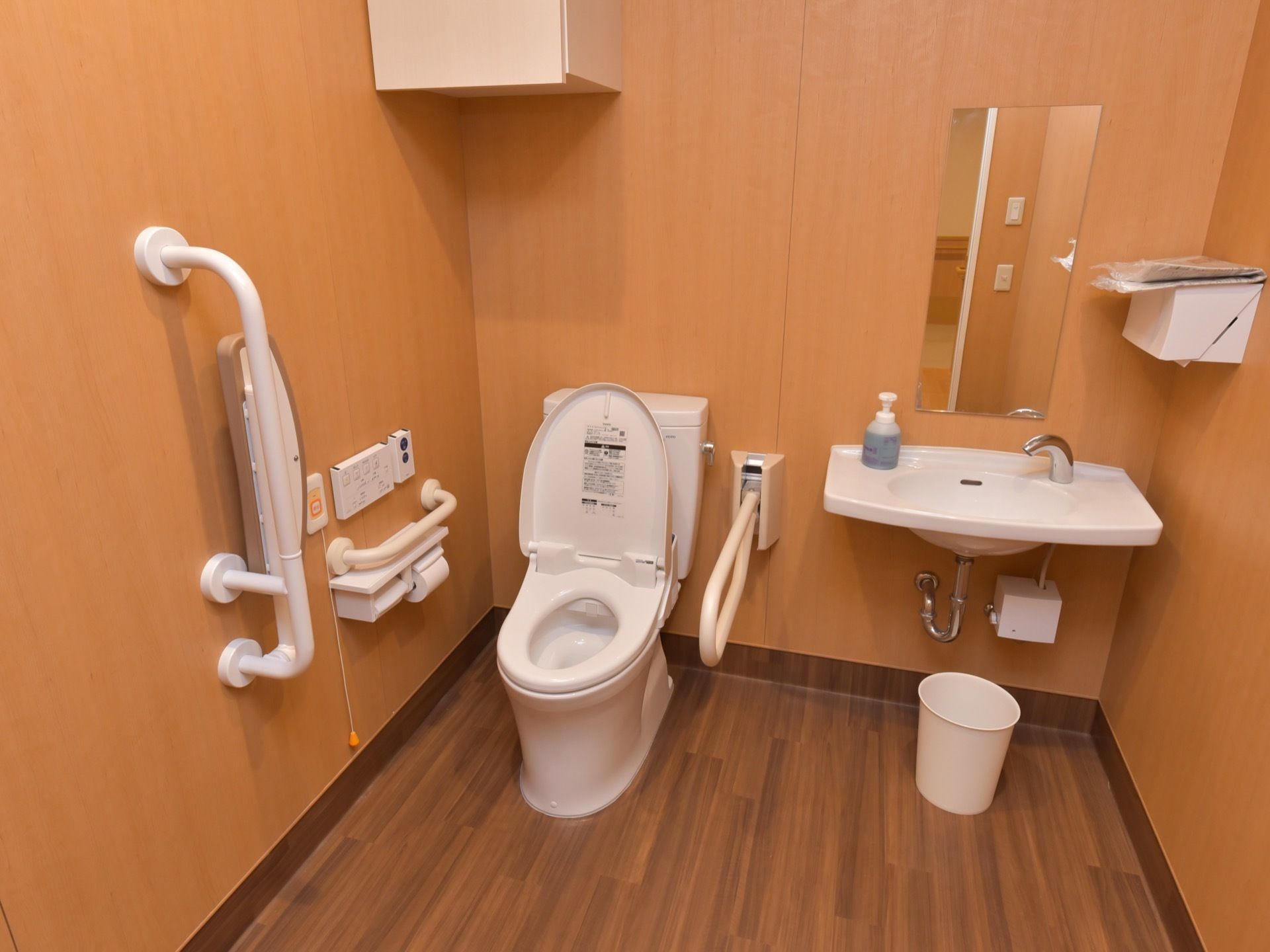 トイレの壁は茶色で、落ちついた雰囲気となっている。直線の手すりや可動式の手すりが設置されている。隣りには洗面台もある。