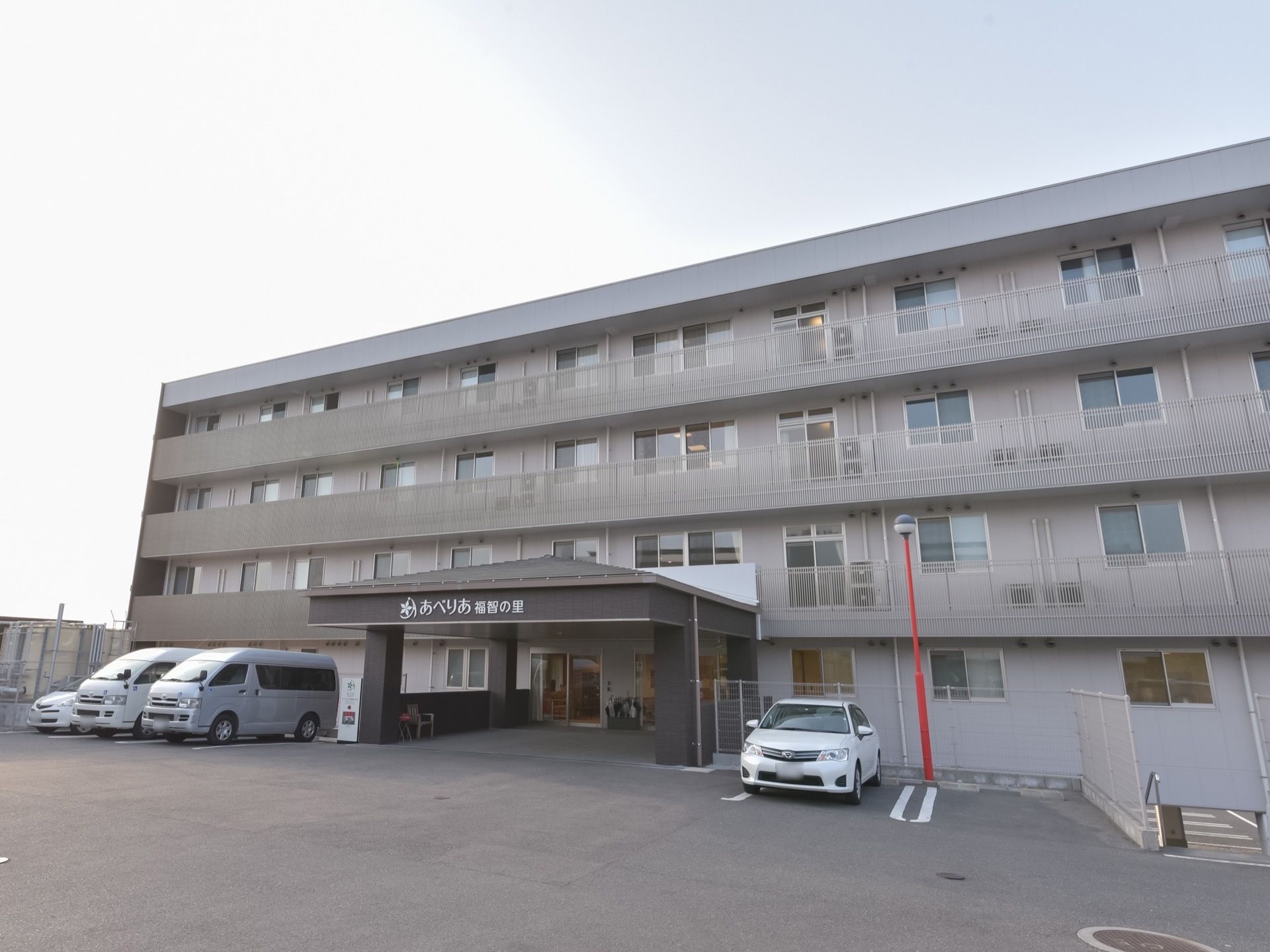 グレーの建物に、ダークグレーの玄関ポーチが飛び出している。玄関前の屋根に施設名が表示されている。玄関周りには駐車スペースがある。