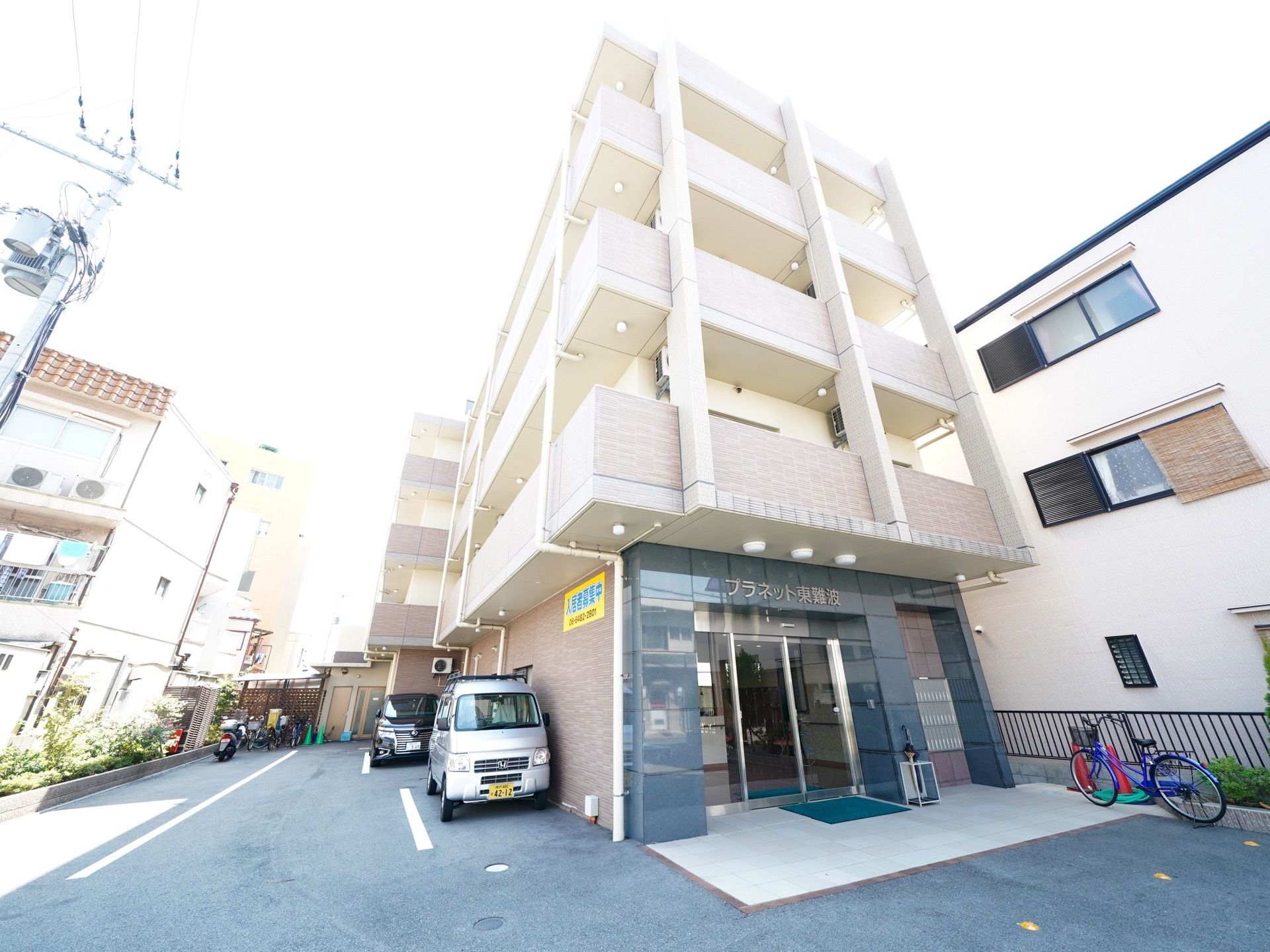 施設は4階建てで、白色とベージュの外壁が使われており、明るくなっている。入口横のスペースには車やバイクが止められている。