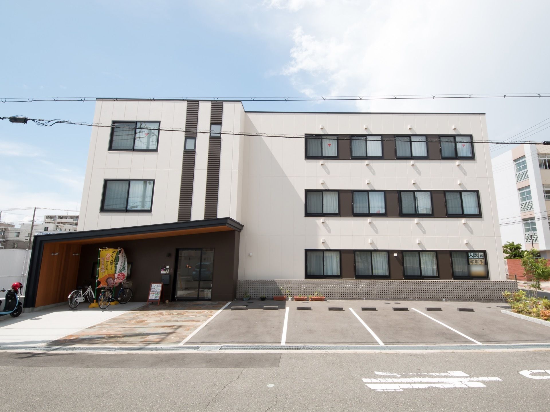白い四角い３階建ての建物の前に４台分の駐車スペースがある。１階の窓に入居者募集中と張り紙が貼られている。