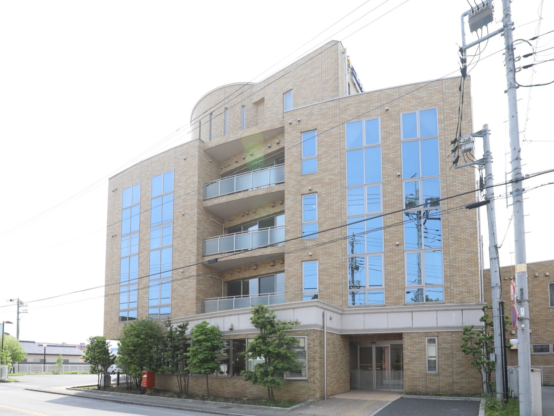 大きな窓とレンガ調の外壁が印象的な５階建ての建物。道路脇に建っているので、１階部分の周りには背の高い木が植えられている。