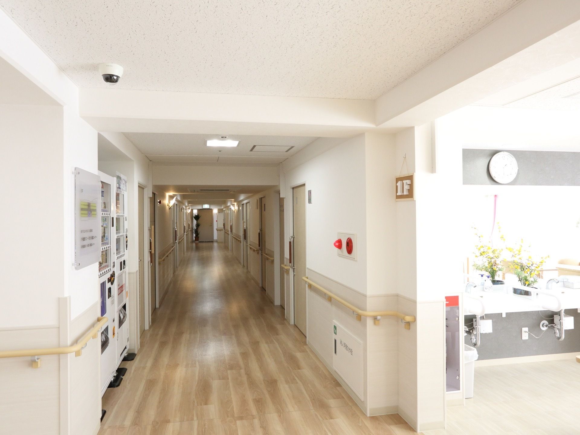 木目調の床の通路に、居室が並んでいる。自動販売機が設置されていて、壁には手すりが取り付けられている。