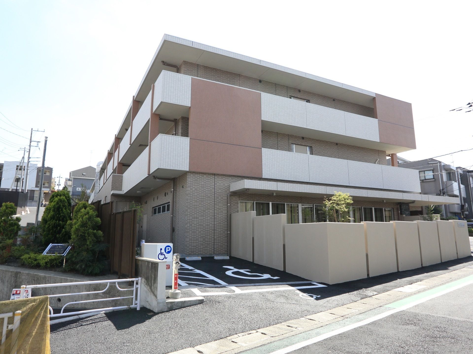 タイルが貼られている。三階建ての細長い建物である。敷地内に白い外壁が造られていて、中の木が見えている。