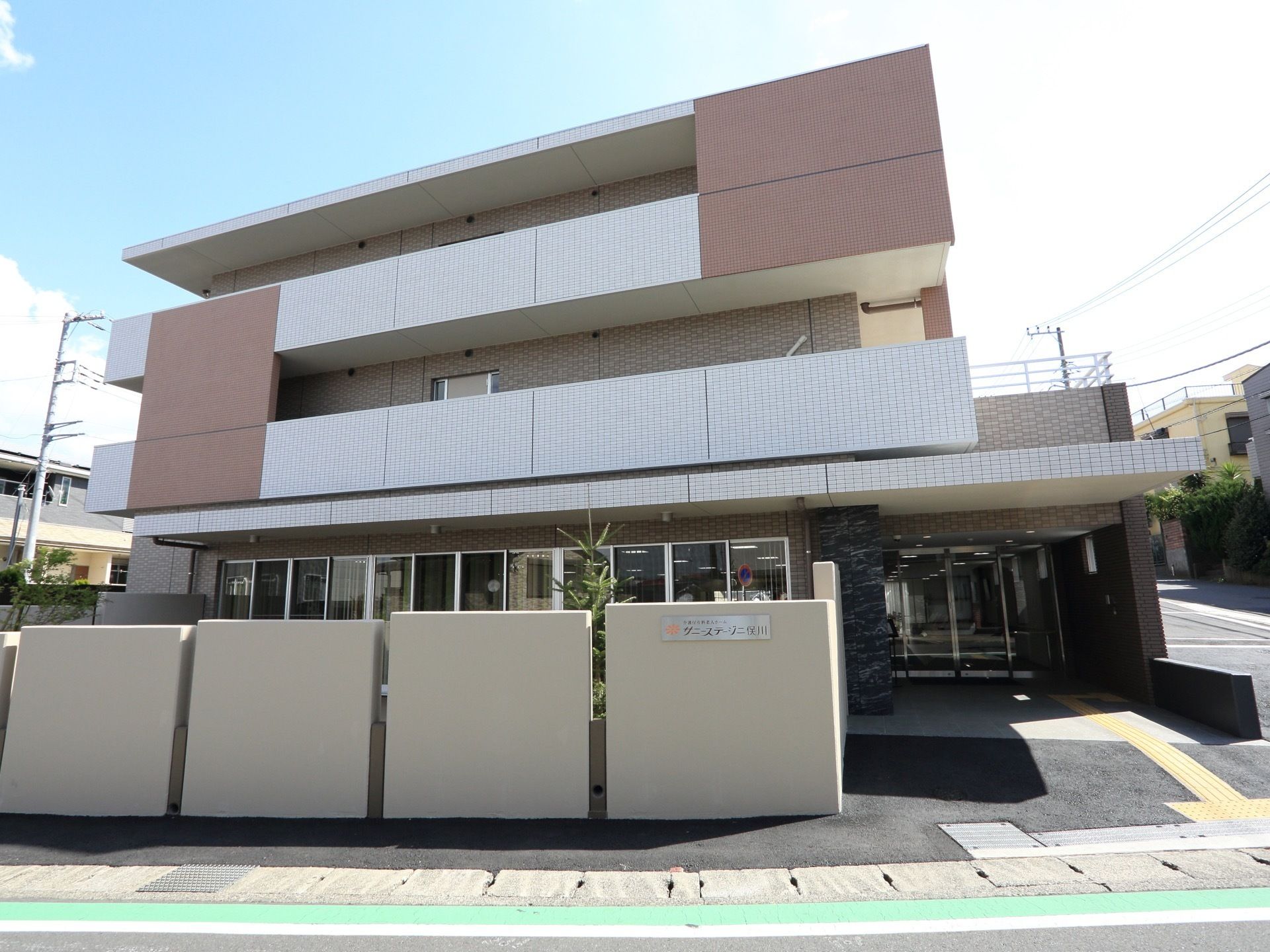 白と茶色のタイル貼りの建物が建っている。少しずつ間隔をあけて白い塀が建っている。その横から自動ドアの玄関が見えている。
