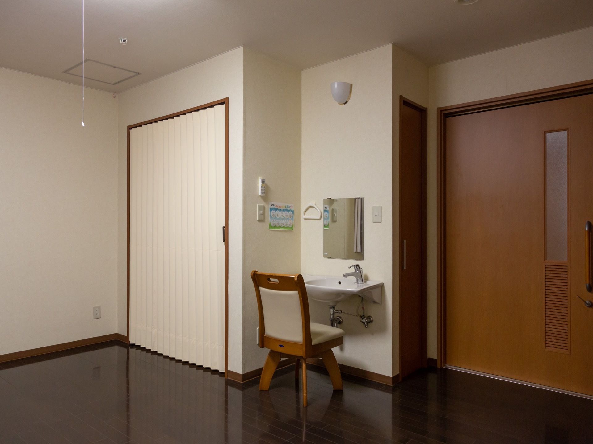 居室の扉の隣には収納スペースの扉がある。壁に洗面台が作りつけられ、その前に椅子が１脚置かれている。天井近くに照明がある。