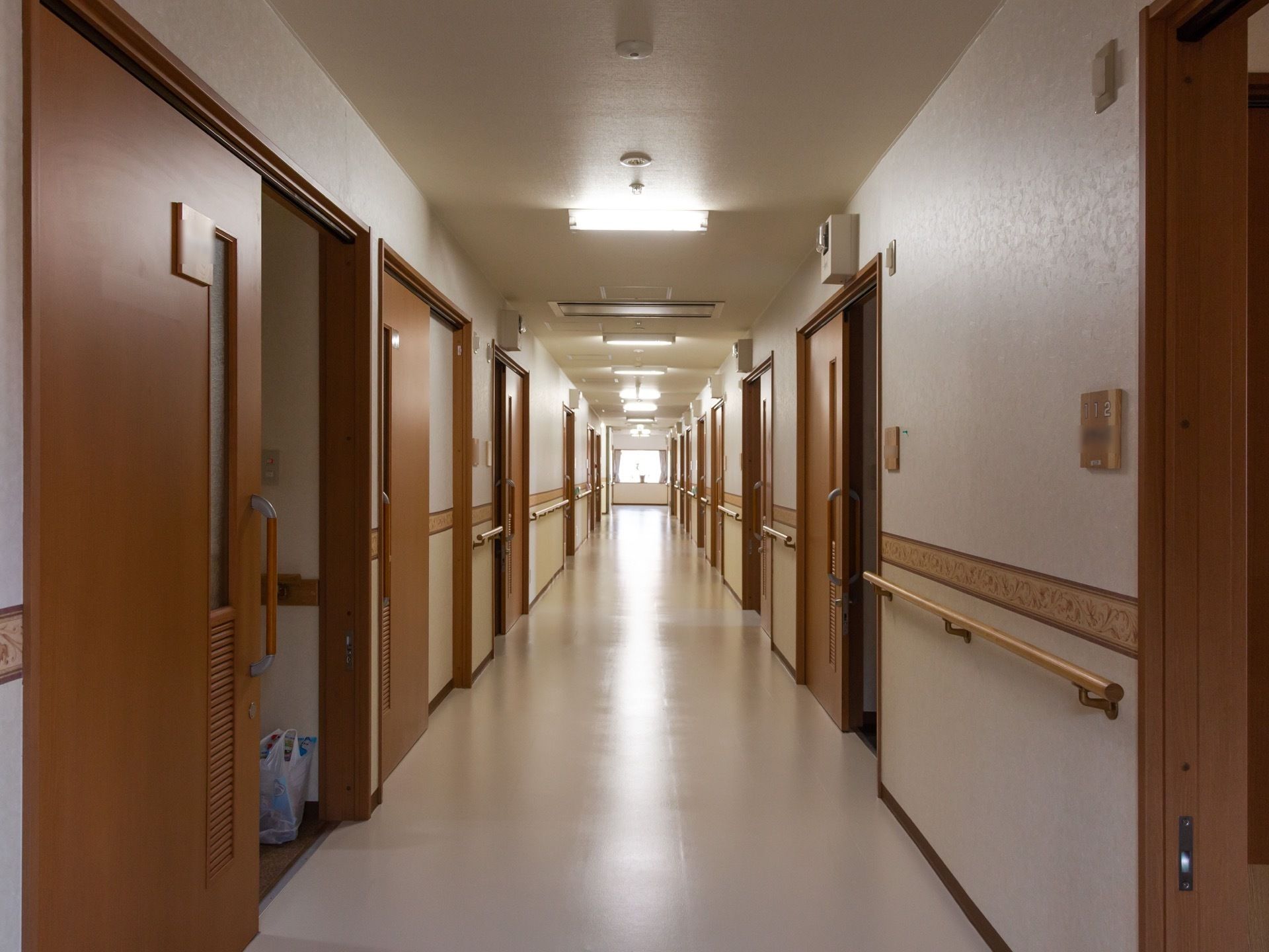 廊下の両側に居室などの扉が並んでいる。突き当りの窓から外光が差し込んでいる。壁に手すりが取りつけられている。
