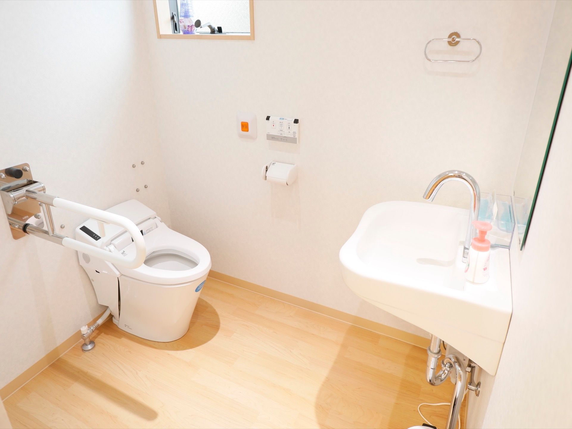 真っ白の洋式便座、洗面台を備え付けたトイレである。可動式の手すりを使いながら、安全に利用することができる。
