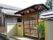 伝統的な日本家屋のような数寄屋門があり、その先に瓦屋根の建物がある。門の前には２段の段差があり、門の横には植物がある。