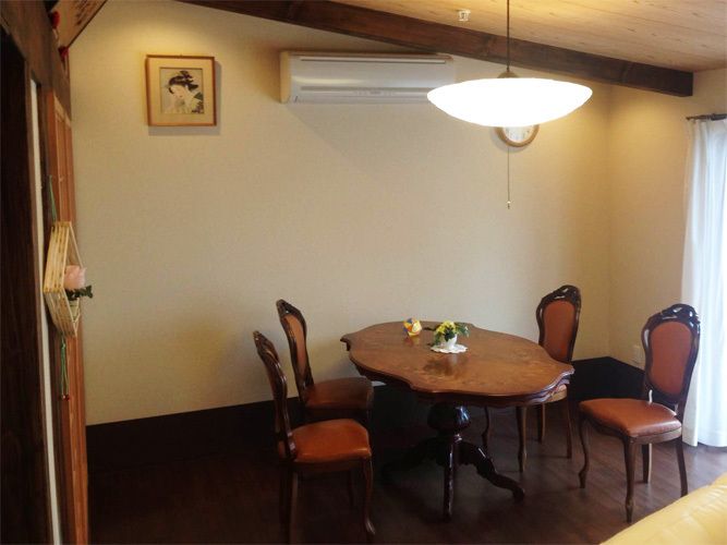 天井から照明が吊り下げられている部屋。その下にはテーブルと４脚のイスがある。壁にはエアコンの他、小さな美人画が飾られている。