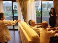 入居者様とスタッフが両手を前に伸ばして体操をしている。3人はソファに座っており、1人はソファの後ろに立っている。
