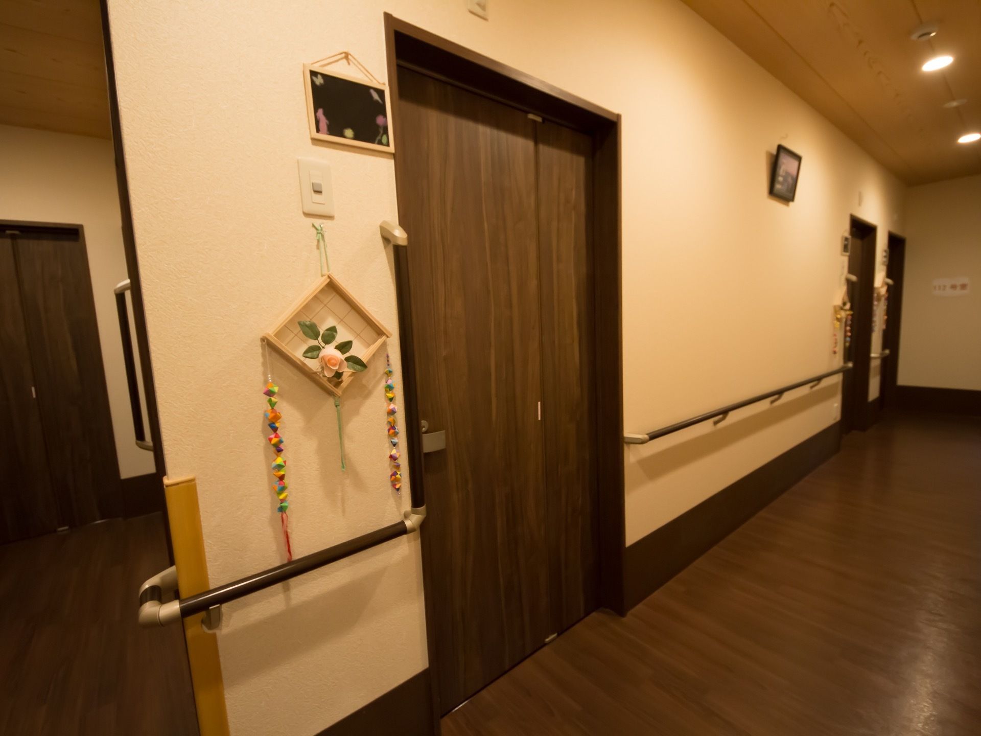 居室前の廊下の床はダークブラウンを使用している。壁には手すりが取り付けられている。居室の扉は幅が広いタイプになっている。