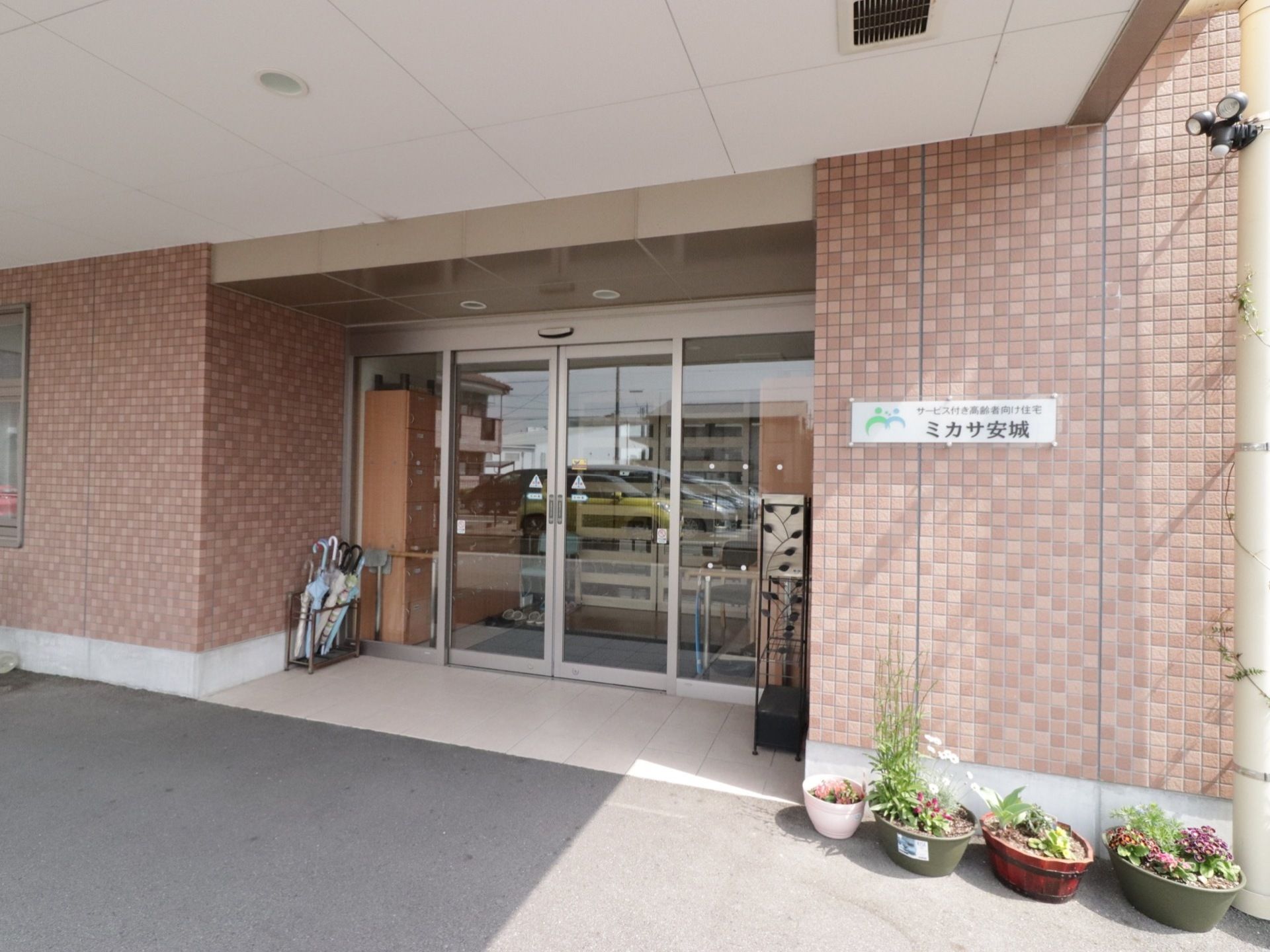 玄関前は広く屋根がある。自動ドアの隣に傘立てがあり植木鉢も置かれている。煉瓦調の壁には施設名が入った表札がある。