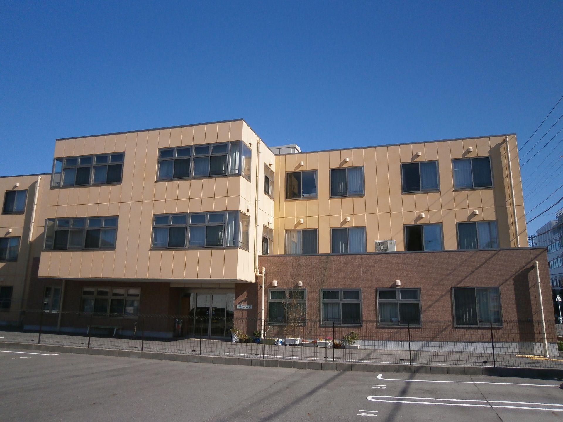 地上３階建て。ベージュとブラウンの外壁になっている。施設は道路に面して建てられている。周辺には大きな建物はない。