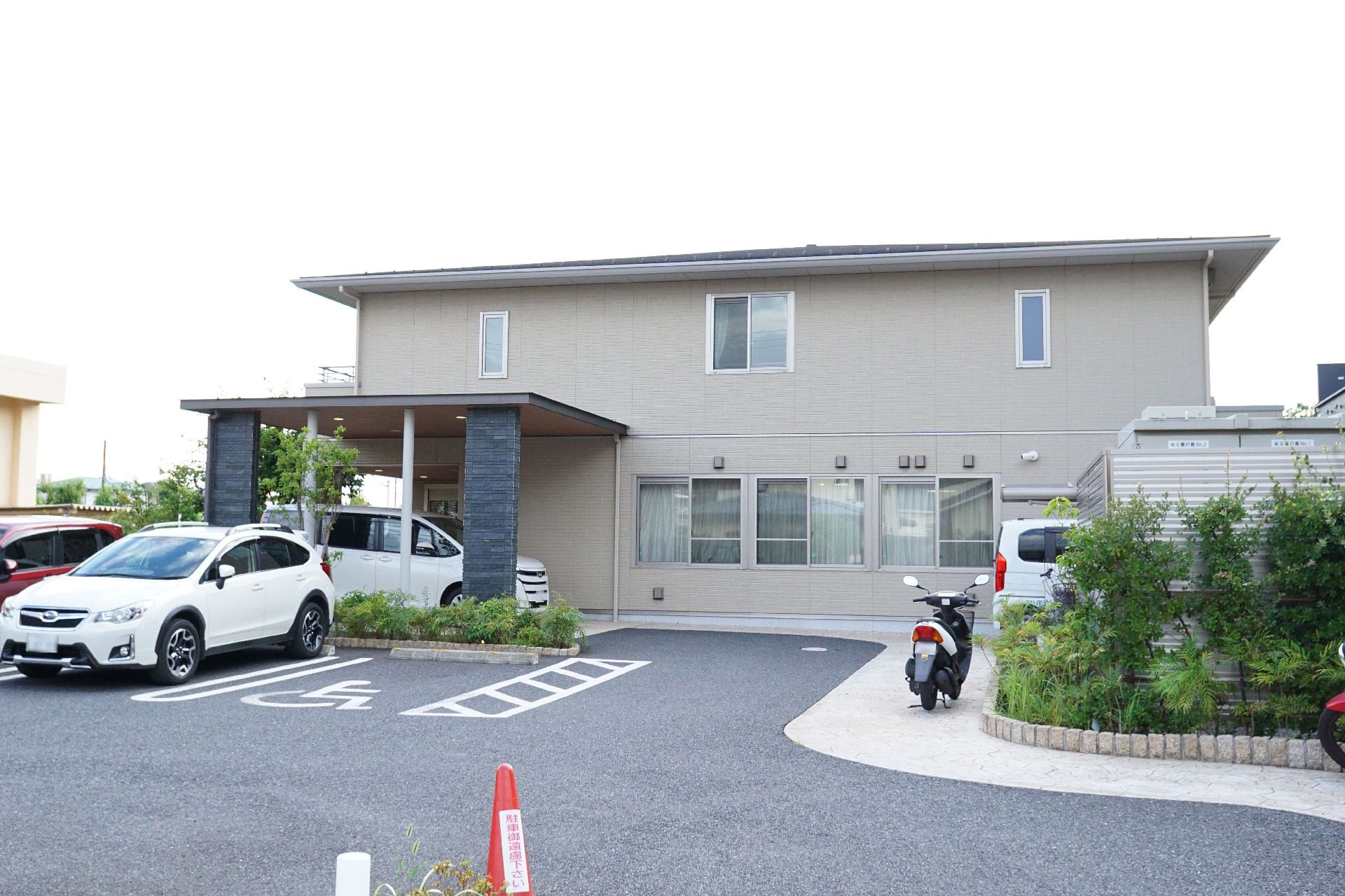 ベージュの外観の２階建ての建物の前にある車が止まった駐車場