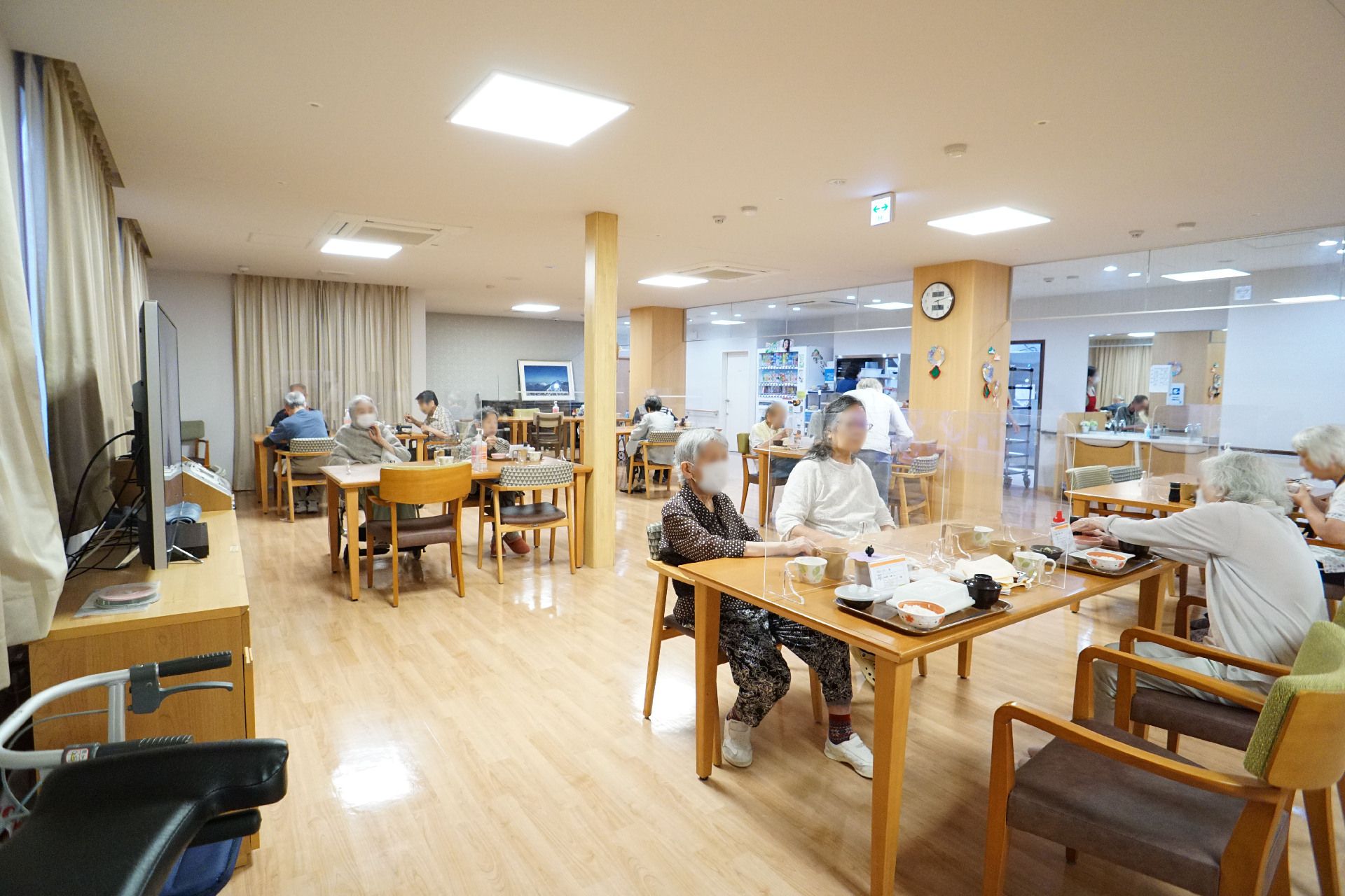柱の立った広間に置かれた透明な仕切りが付いている机の周りの椅子に座る入居者様
