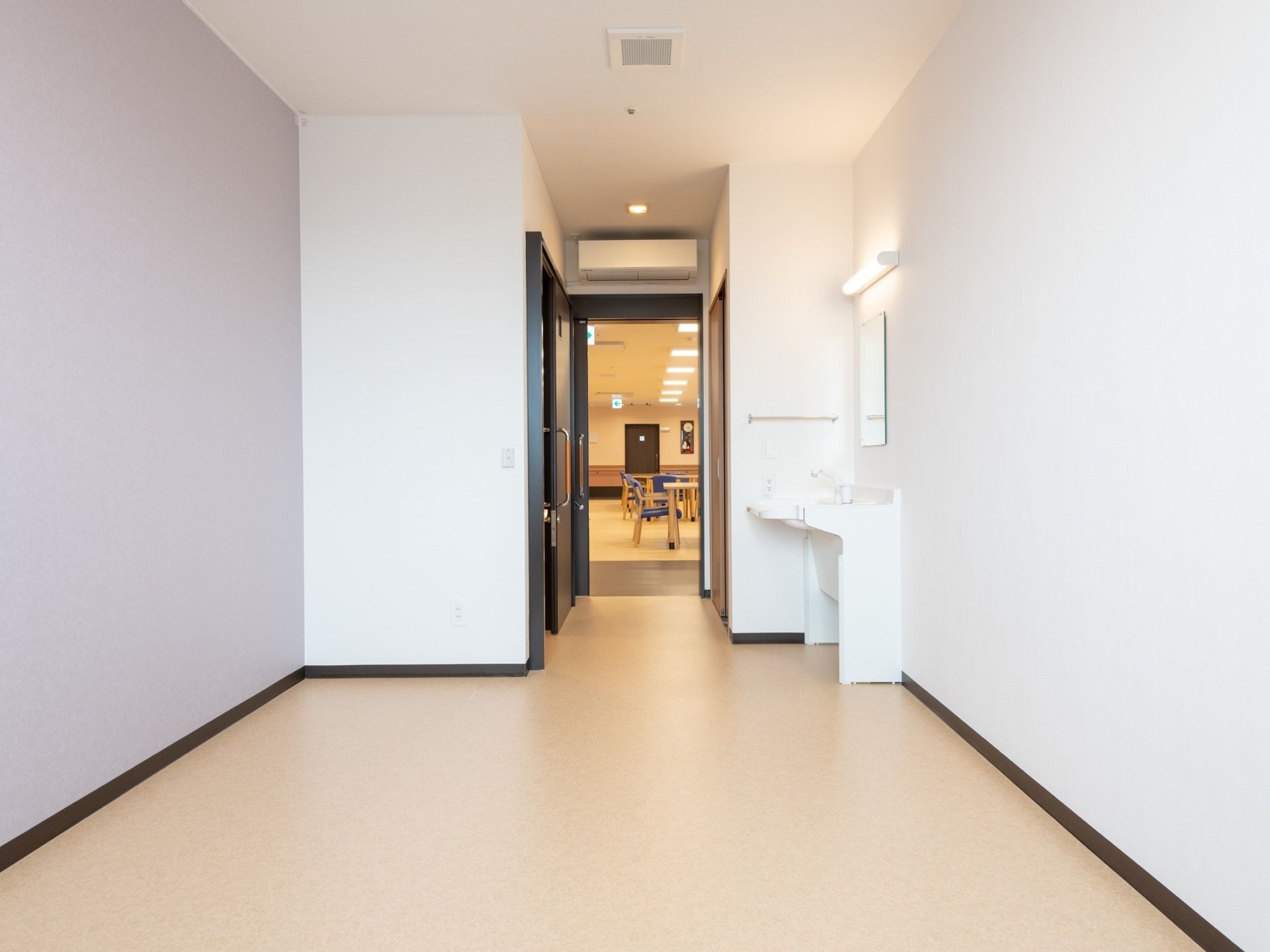 クリーム色の床に、白い壁の部屋がある。壁際には洗面台があり、反対側にはトイレがある。扉があいており、共有のラウンジが見える。