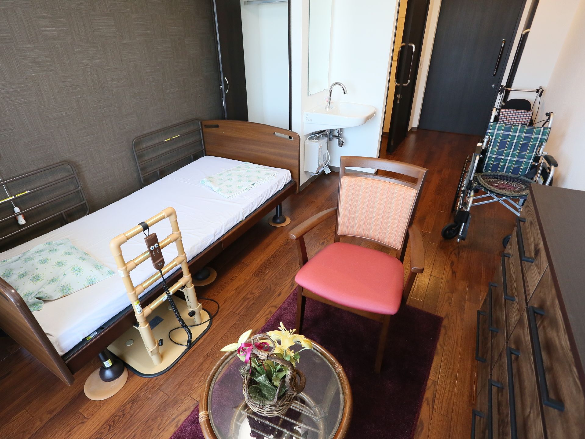 部屋の真ん中に小さな丸テーブルと１人掛けの椅子がある。ベッドには転落防止柵がついており、リモコンが掛けられている。