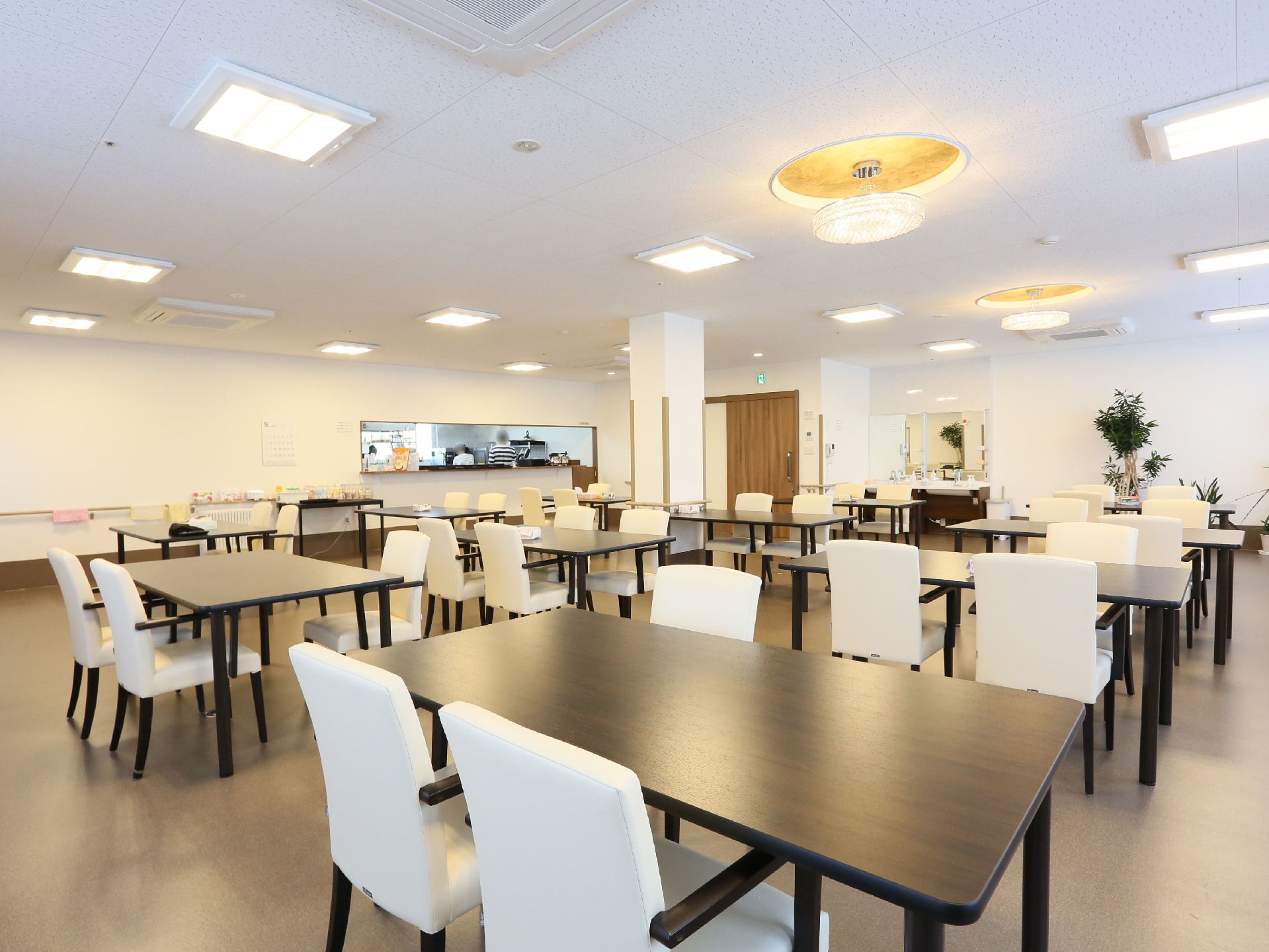 茶色いダイニングテーブルと白い椅子がたくさん並べて置いてある。部屋の角に洗面台があり、壁に鏡が設置されている。
