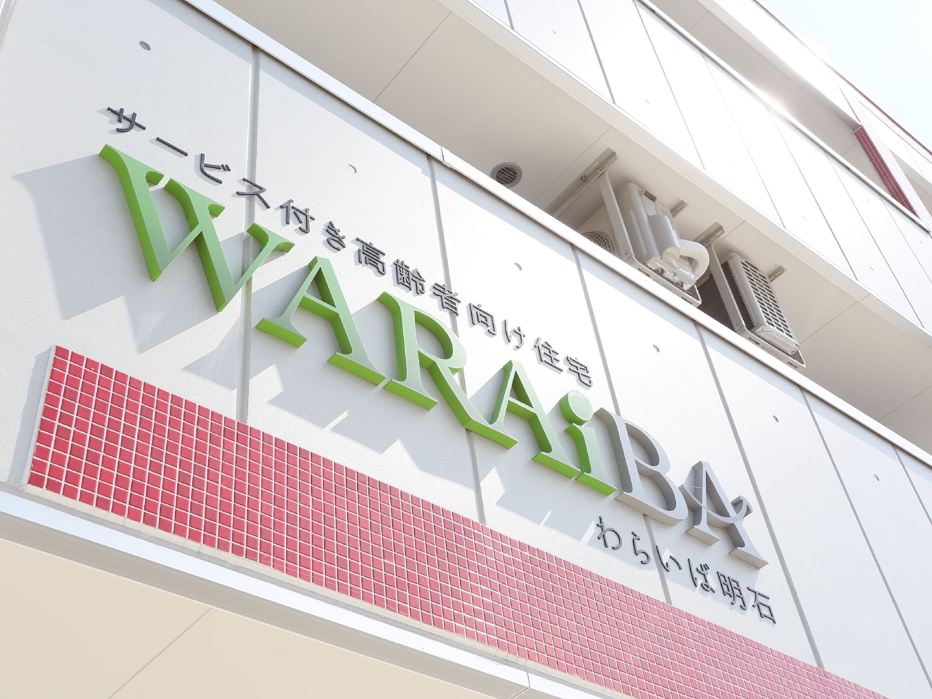 建物の白い外壁に、緑色とグレー色のアルファベットで施設の名称が表示されている。施設名の下は赤の細かいタイルで装飾されている。