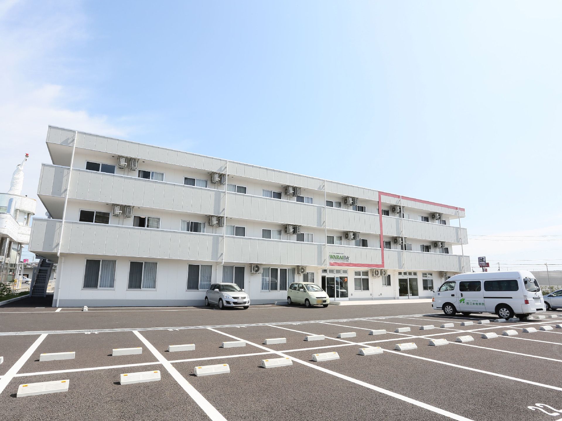 白い３階建てのアパートのような建物がある。アスファルトで整備された広々とした駐車場に、数台の車が停められている。