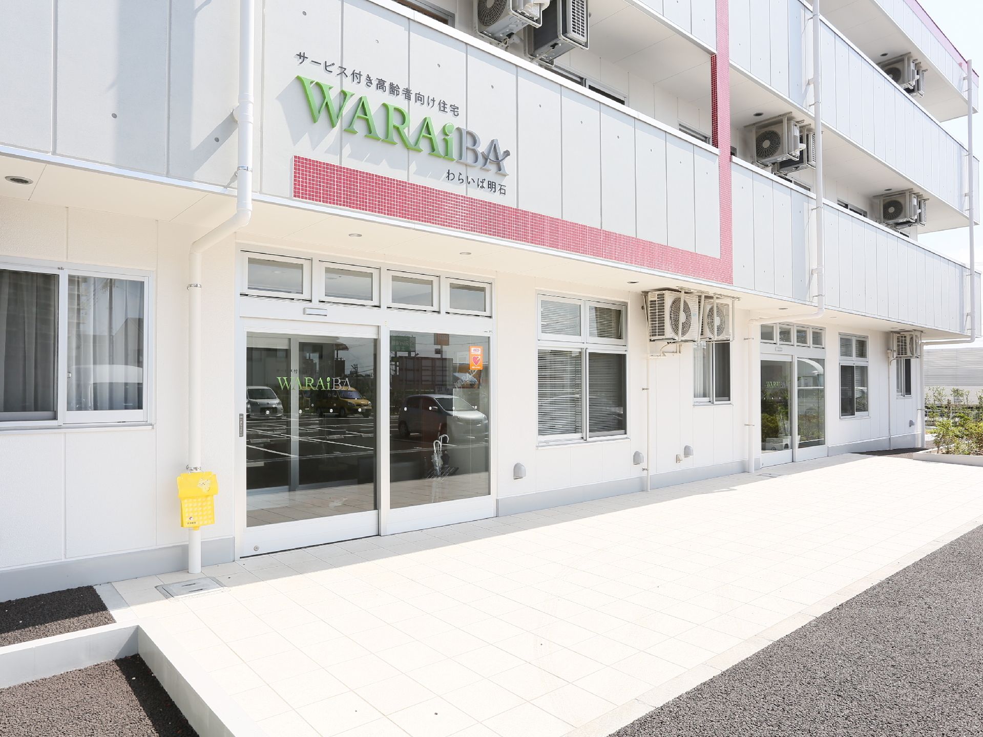 マンションのような大きな建物である。１階の側面の窓は２階に届くほどの高さがある。建物の周りには植栽がある。