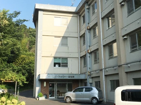 ４階建ての建物の前には駐車場があり、周辺には緑が多い。