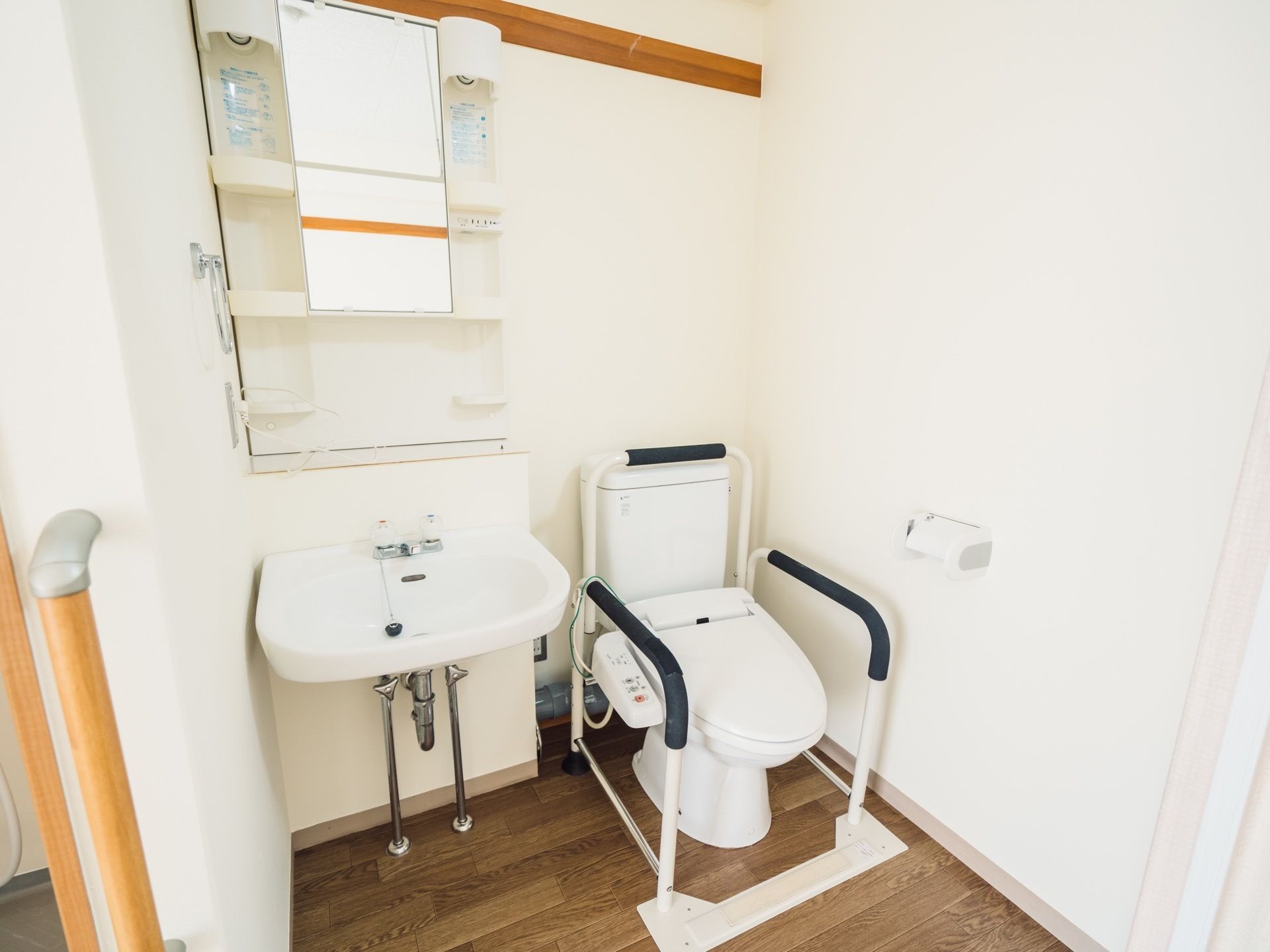 白と木目を基調としたトイレスペースである。便座には、背もたれと肘掛けを備えている。横側に洗面台を設置している。