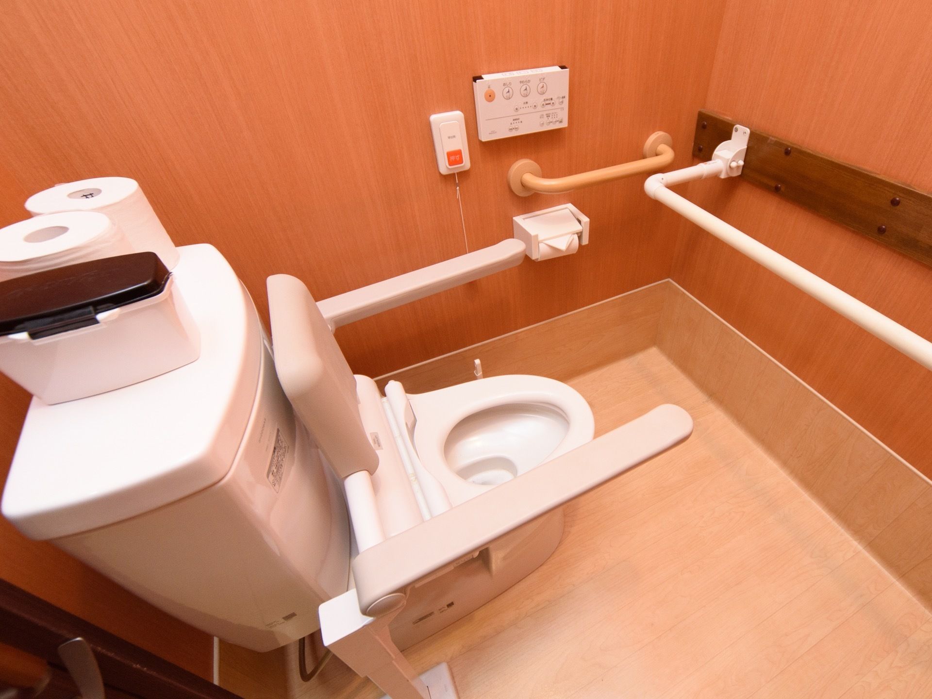 淡い木目調の床と濃い木目調の壁を組み合わせたトイレである。温水洗浄便座が設置され、手すりが多個所に取り付けられている。