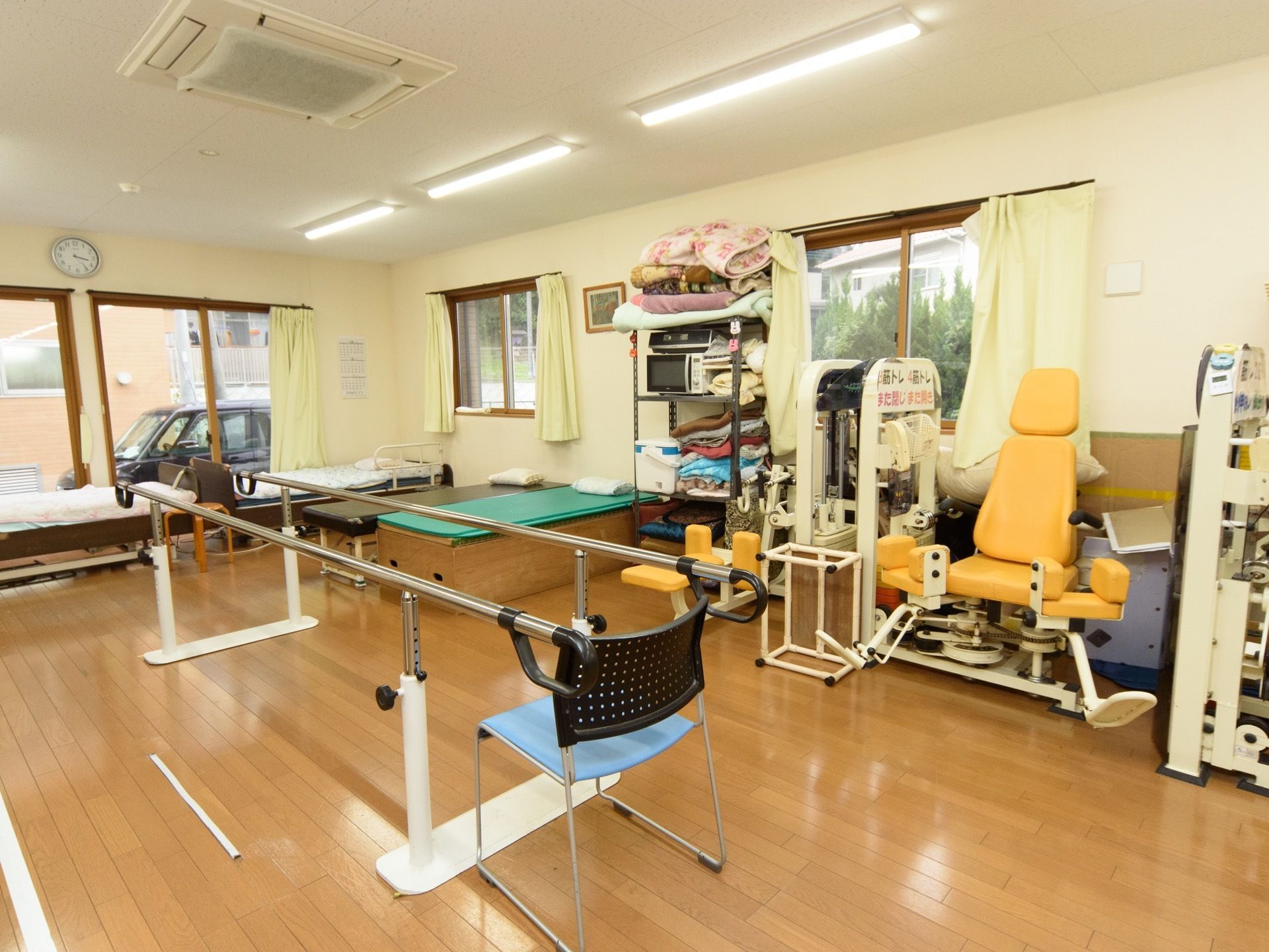 機能訓練室には、歩行用の平行棒や筋トレマシーン、ベッドなどが置かれている。ラックにはクーラーボックスや電子レンジ、毛布などがある。
