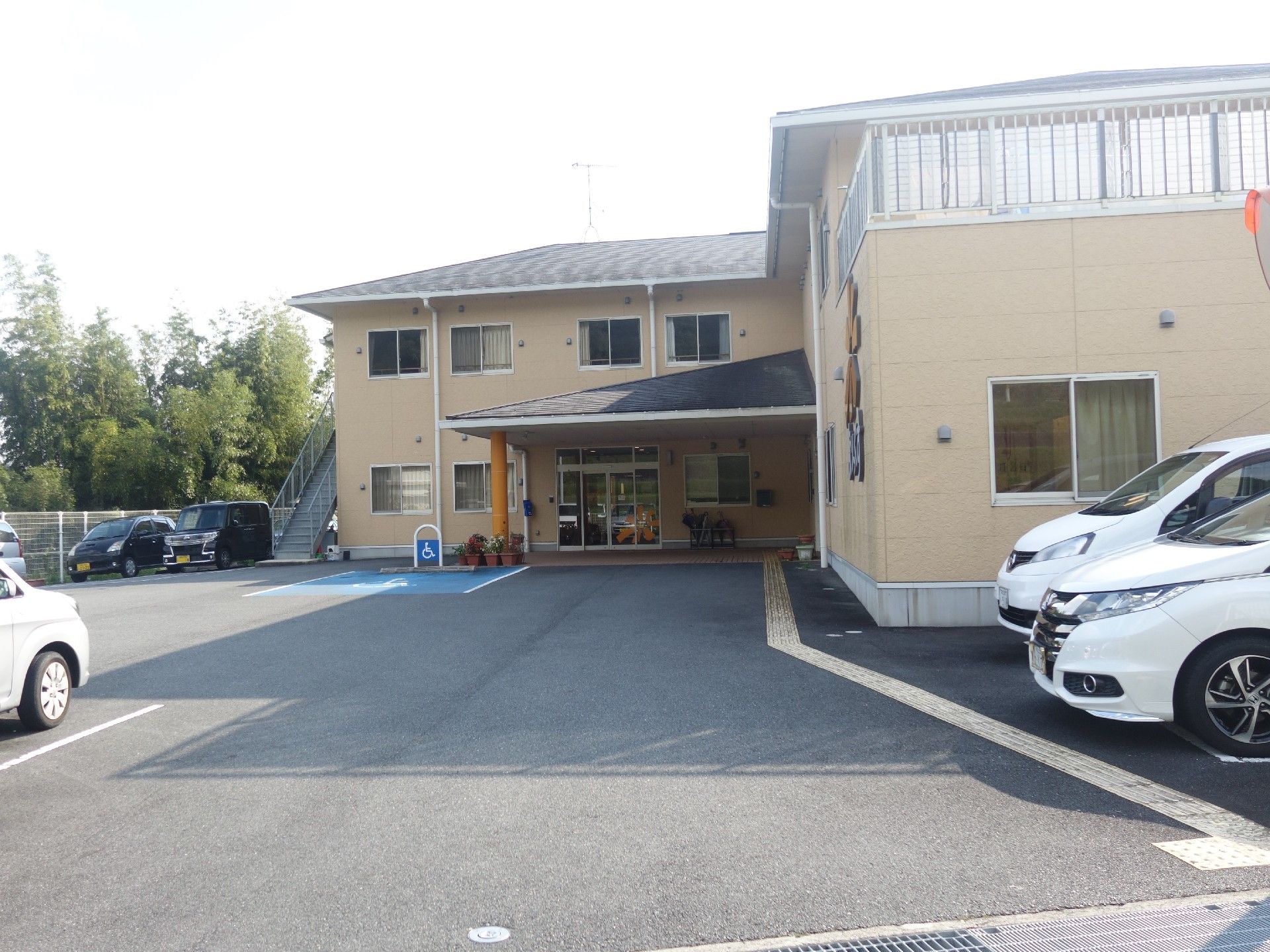 施設の建物の外観である。中央部の玄関には屋根があり、通路はフラットになっている。周囲には駐車場が設置され、緑の木々に囲まれた環境である。