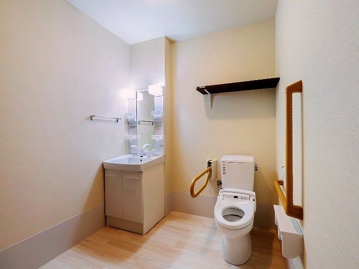 部屋の中にトイレと洗面台がある。トイレは温水洗浄便座付きで左右には手すりがあり、真上には黒い棚がある。