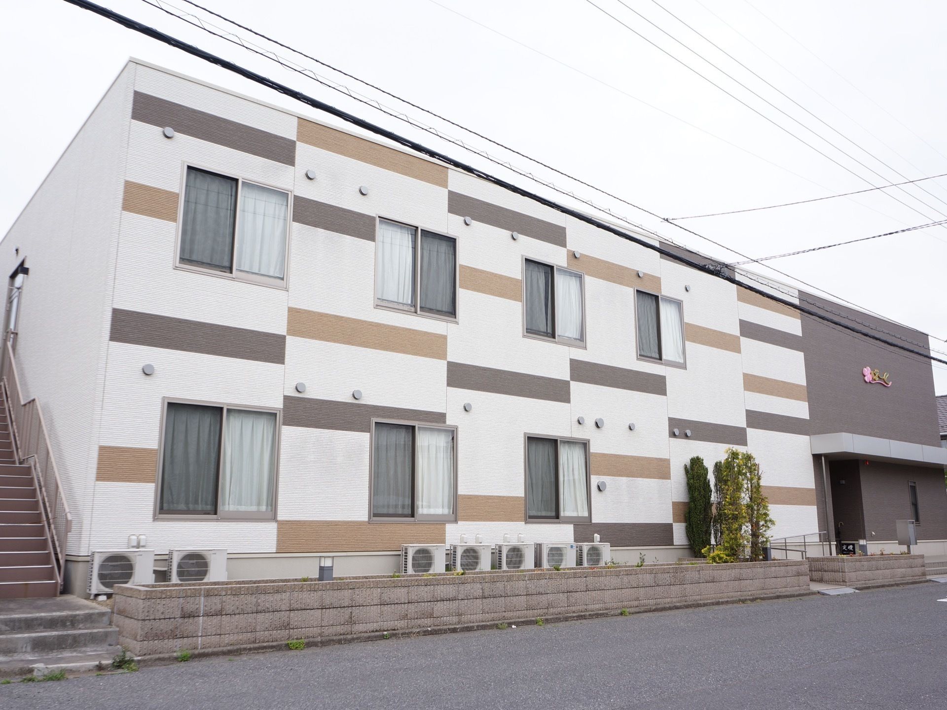 ２階建てのアパートタイプの建物の外装は、白地に変則的に２色の横縞模様がはいって印象的。エントランスのある部分に施設のロゴが付いている。