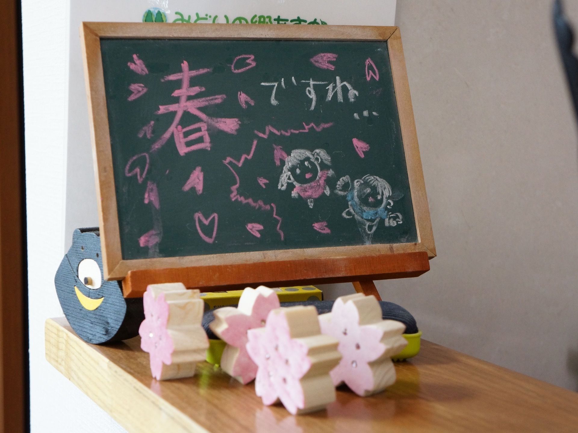 飾り棚に小さな黒板が置かれており、チョークでメッセージが書かれている。木製の熊や桜の置物が飾られている。
