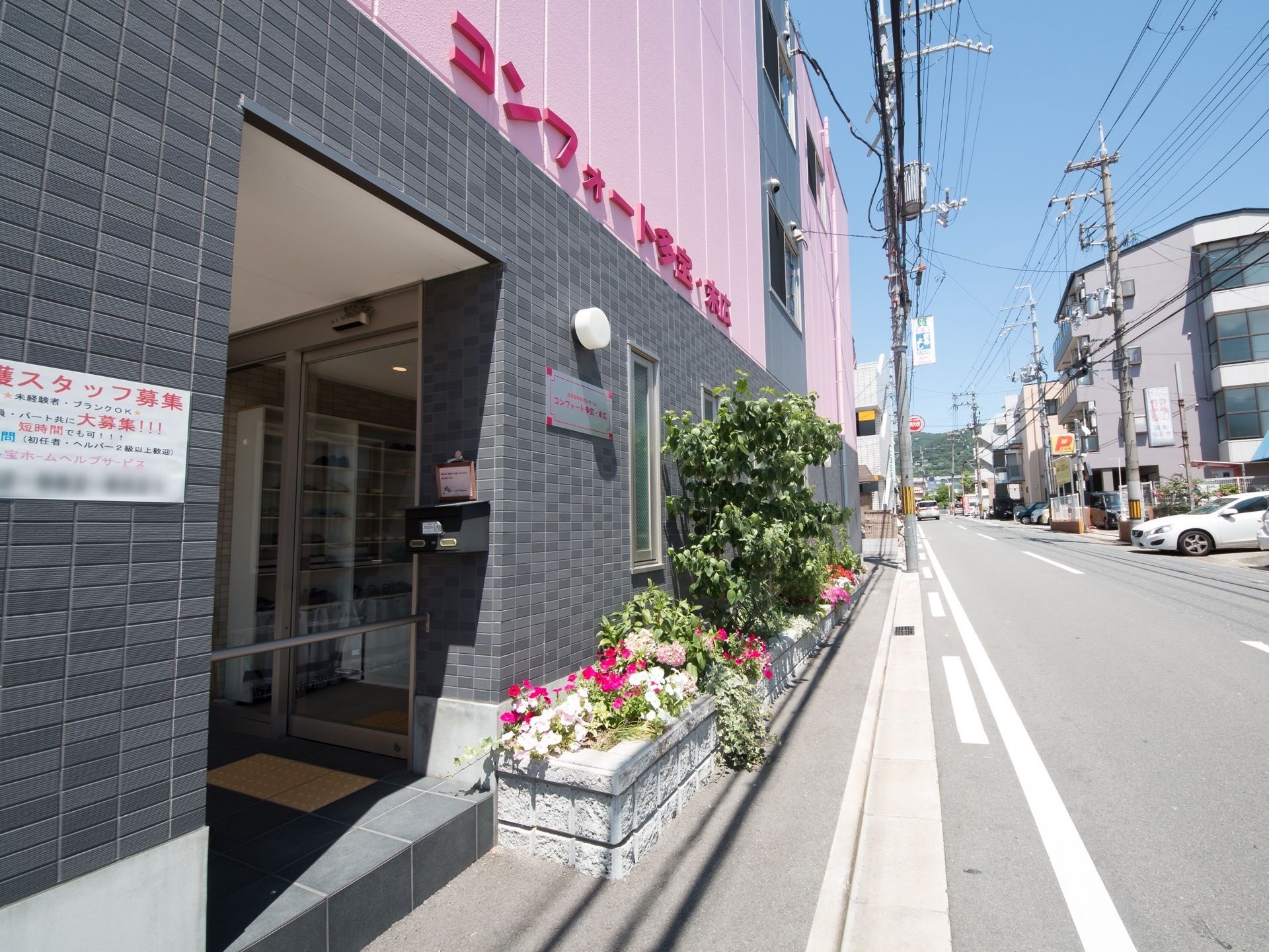 通りに面した玄関の様子である。道路脇に色とりどりの花や木が育てられている植込みがある。1階外壁はシックなグレーのタイル張りである。