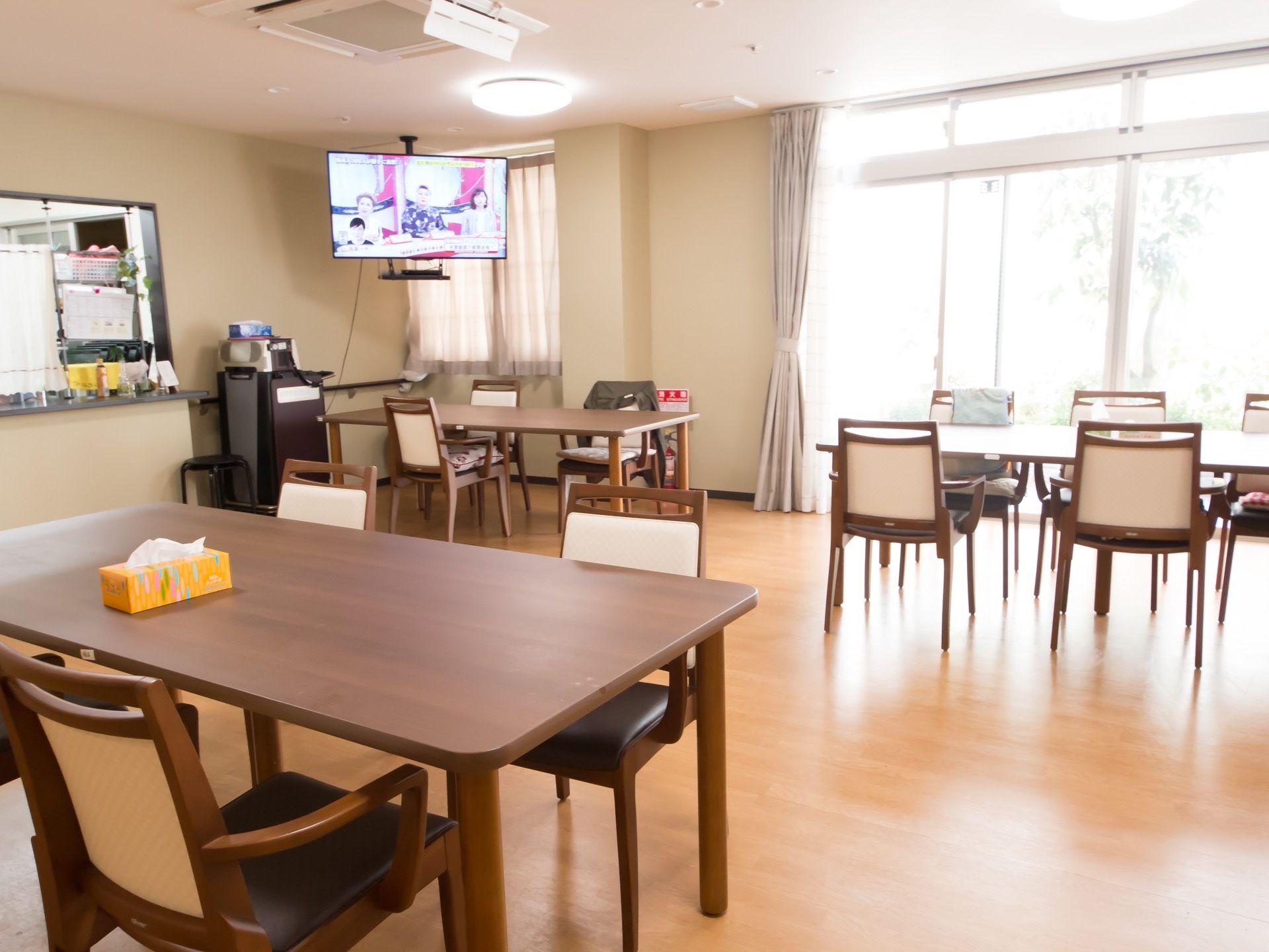 数組のダイニングテーブルと椅子のセットが置かれている。奥には厨房がある。天井からテレビが吊るされている。