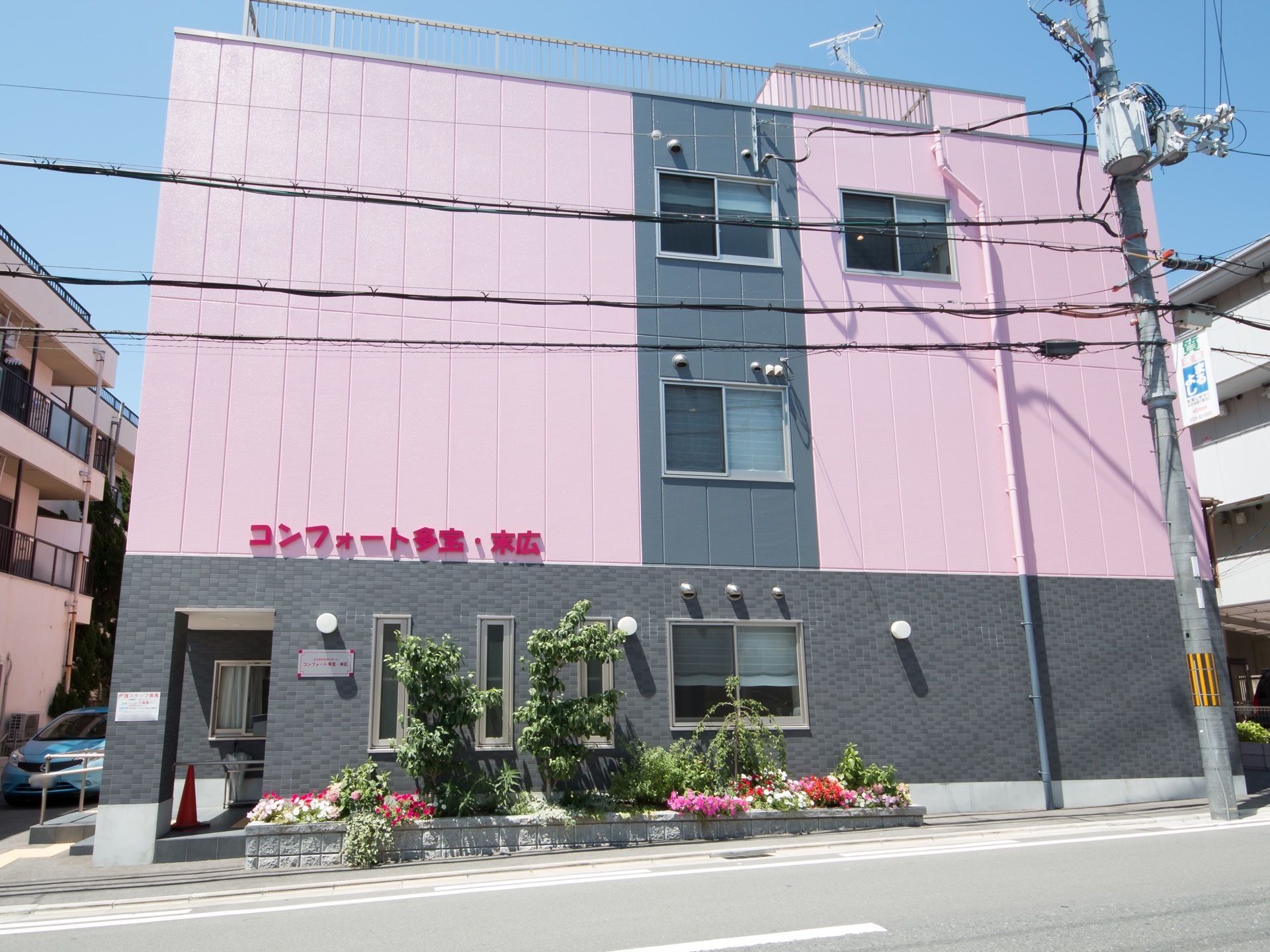 ピンク色の目立つ３階建ての建物となっている。建物の２階部分の壁にに大きく赤い文字で施設名が入れてある。施設の横に花壇が設けてある。