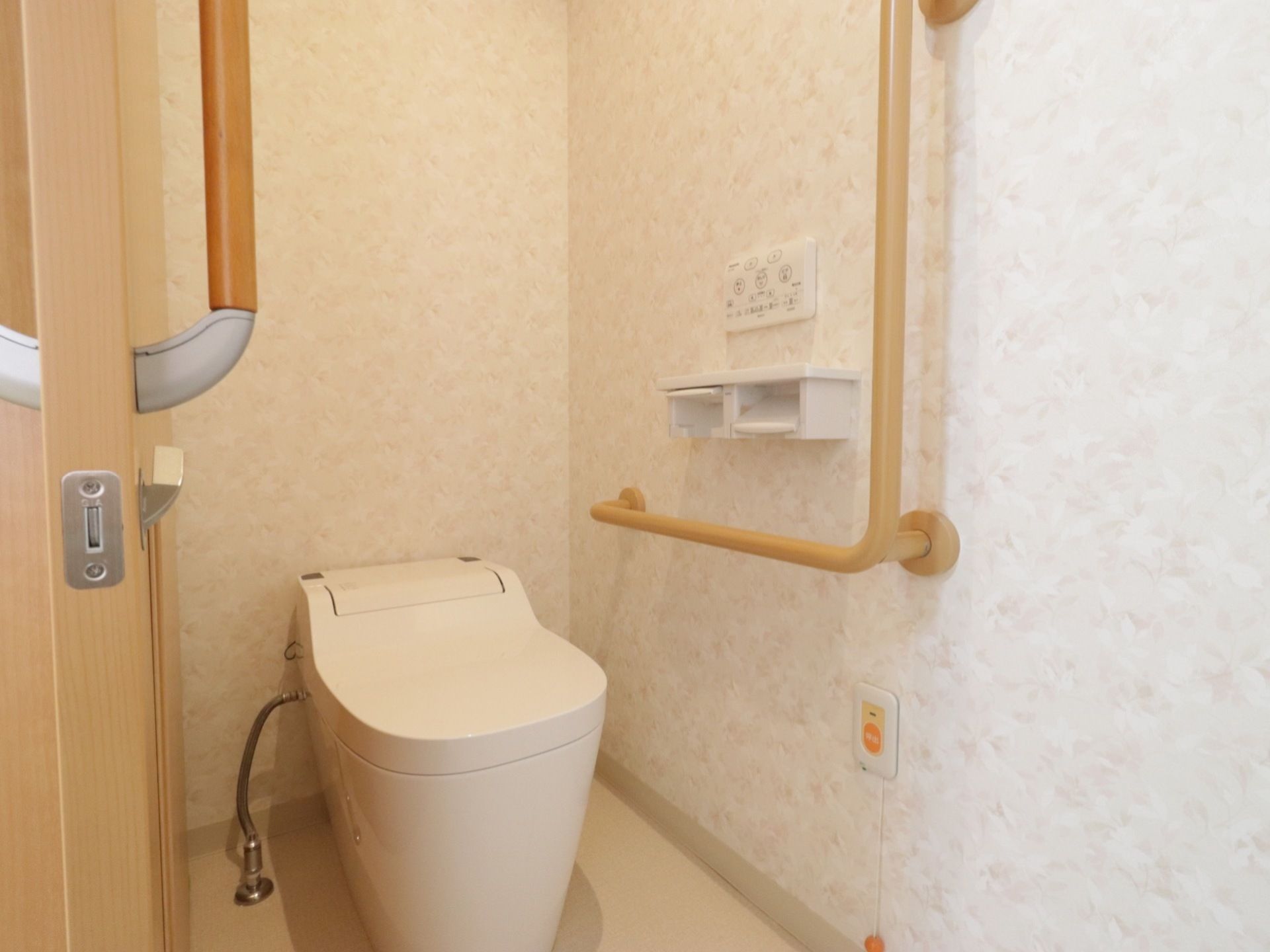 模様入りの壁のトイレスペースである。横側に引き戸のドアがあり、壁際にエル型の手すり、ナースコールを設置している。
