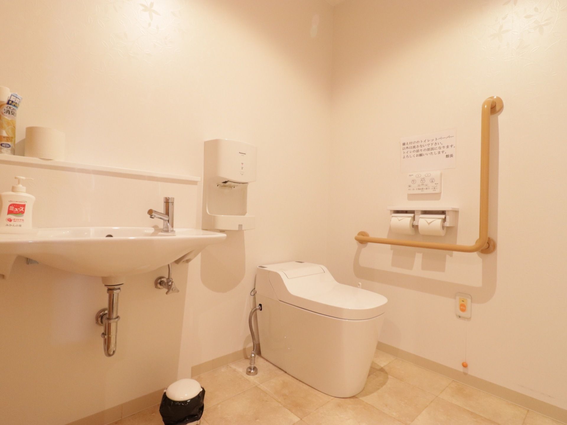 やわらかい白を基調としたトイレスペースである。壁にエル型の手すりとナースコールなどを備えている。横に洗面台を設けている。