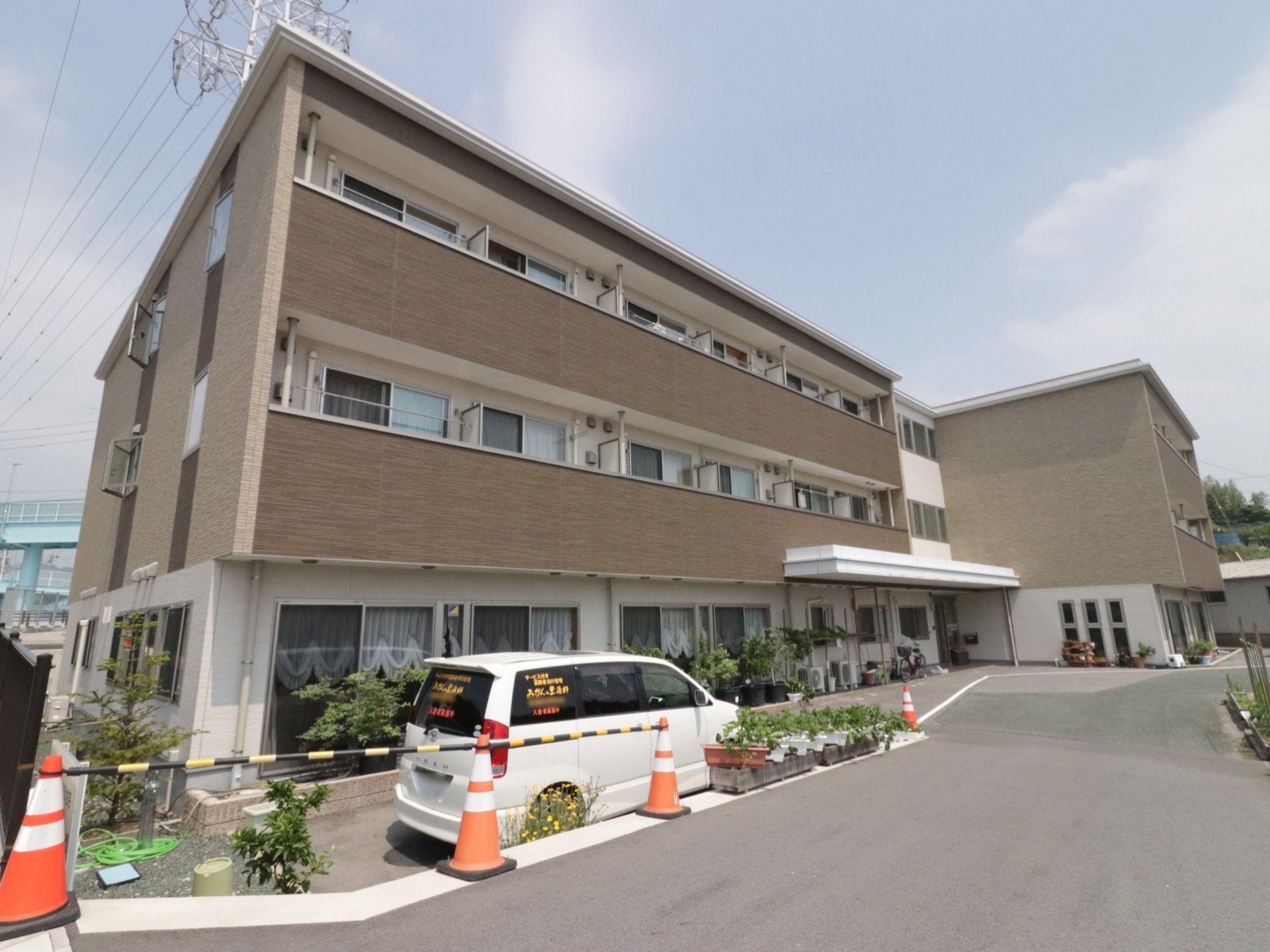 エル型構造の３階建ての建物である。建物前に、白の施設専用の車がある。敷地内出入口の通路の横に緑のプランターを飾っている。