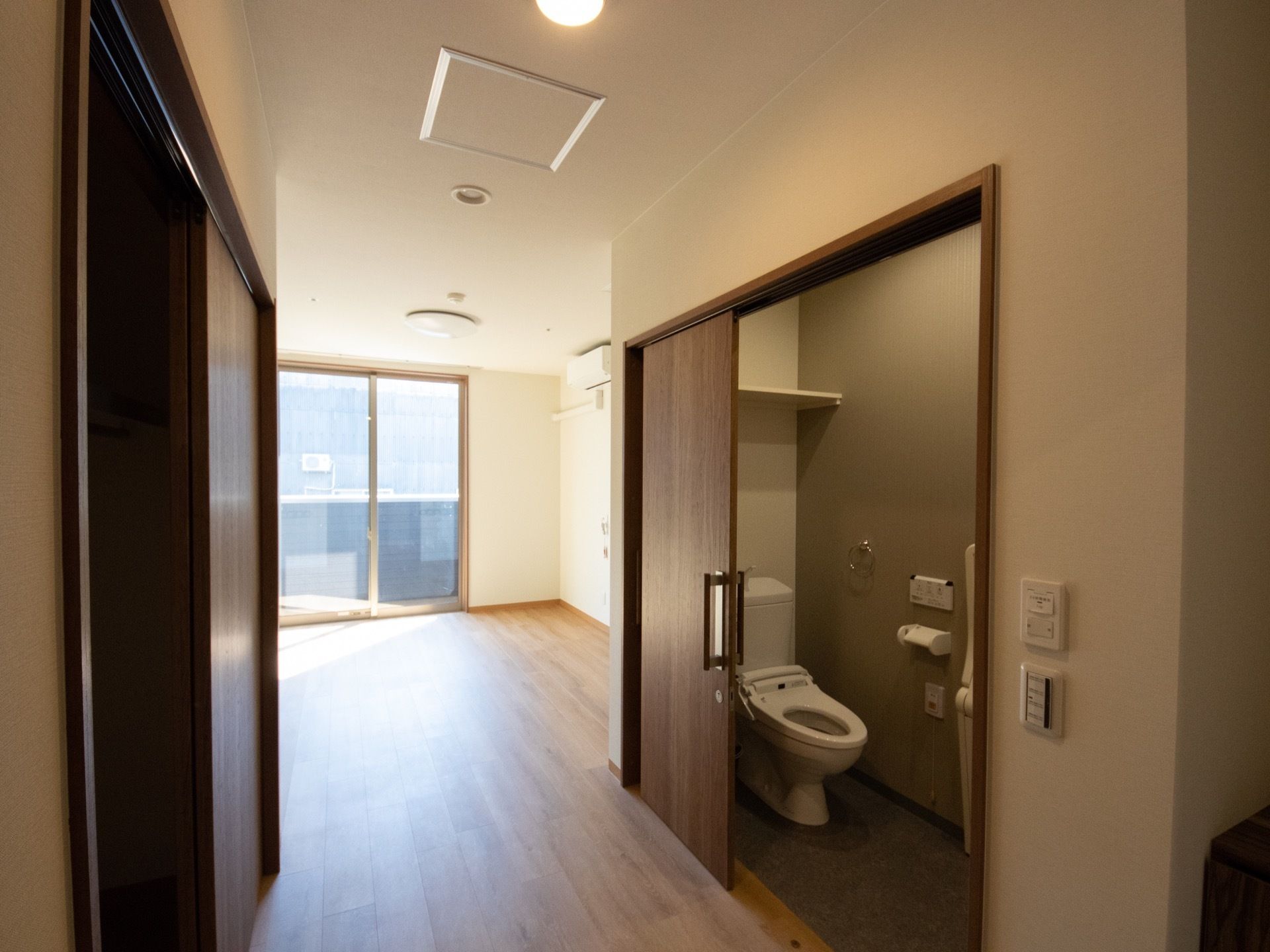 居室には窓、エアコンがある。茶色い扉のクローゼットの向かいにはトイレもある。トイレタンクの上に収納棚が取り付けられている。