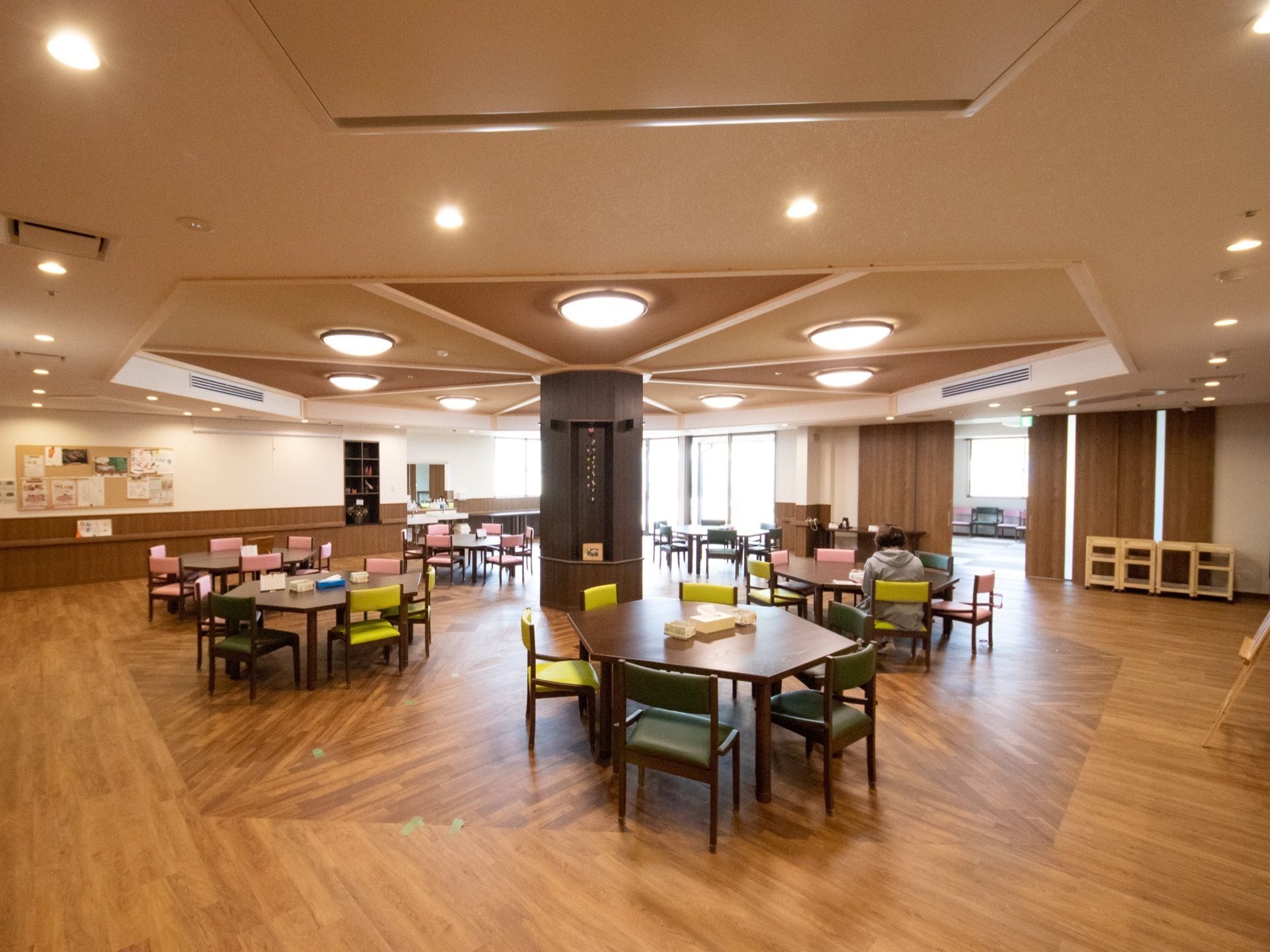 明るいリビング。壁には掲示板があり、行事のお知らせなどが貼られている。机の上にはカレンダーや小物入れが置かれている。