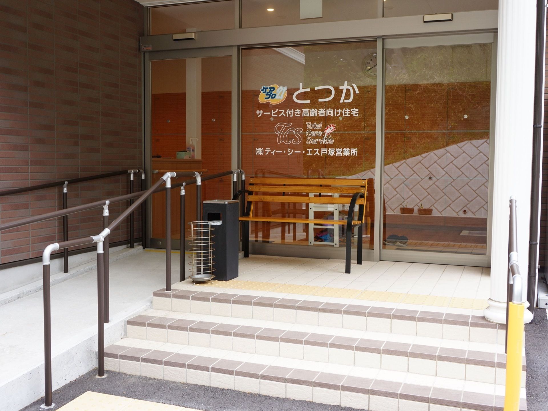 玄関ドアはガラス張りで自動ドアになっている。ドアに施設名が表示されている。3段の階段と隣に緩やかなスロープがある。