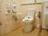洋式の水洗トイレが設置されている。便座には可動式の手すりがつけられている。便座の右側に洗面台があり、緊急時コールボタンもある。
